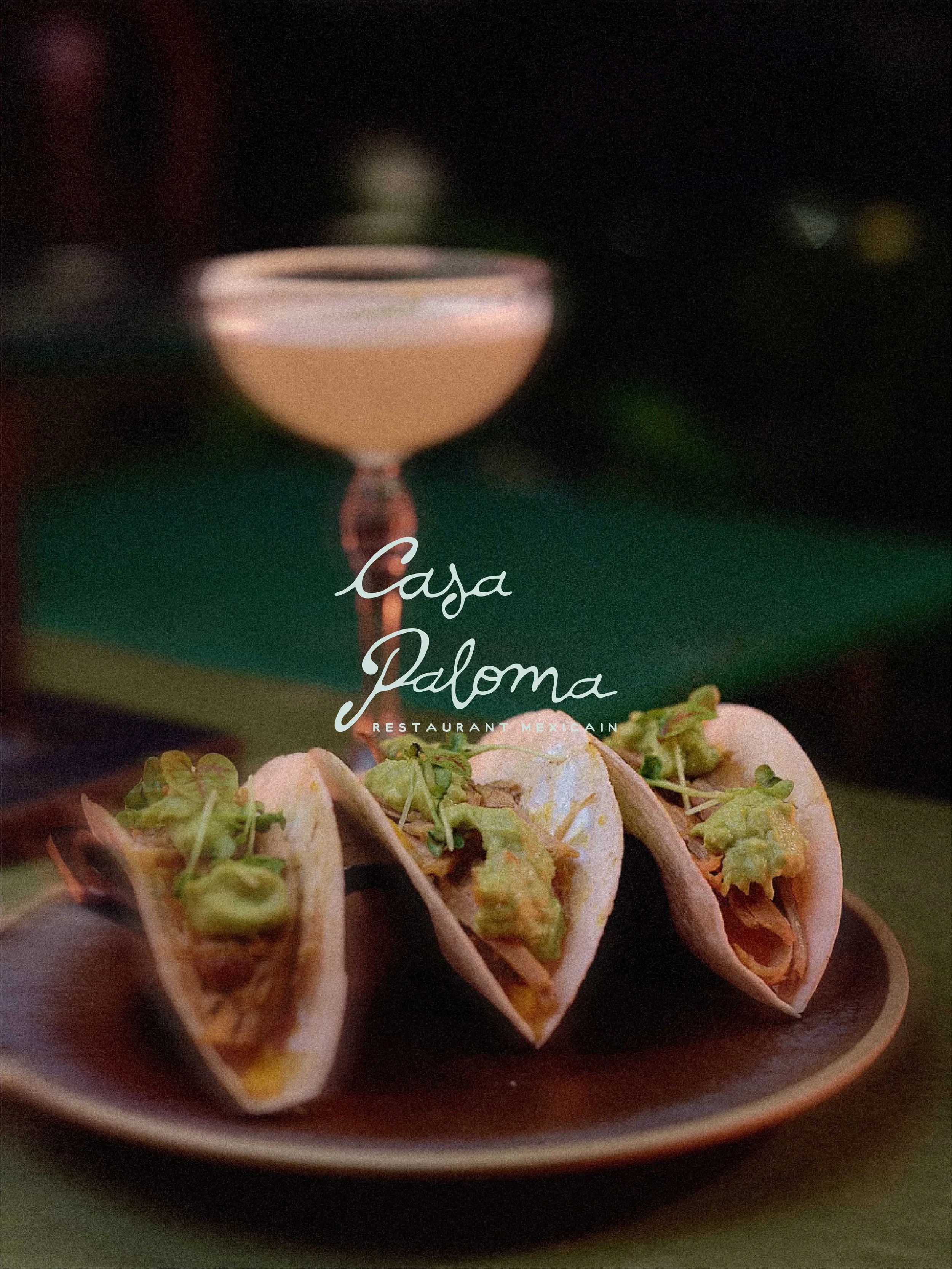 Tacos garnis de viande, guacamole et coriandre sur une assiette en dessous d'un verre de cocktail rose, en arrière-plan flou.