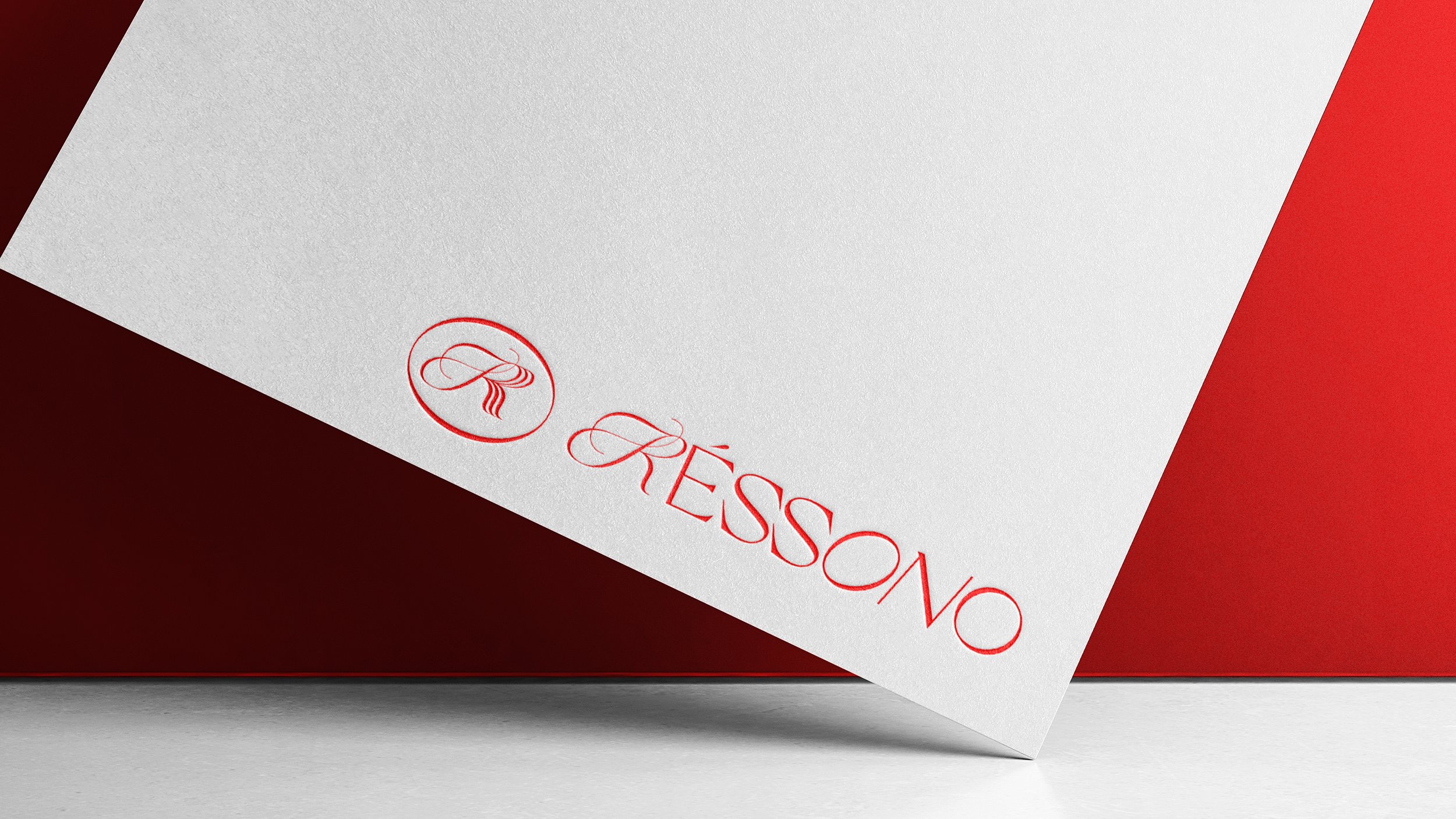 Close-up of a white card with red text and logo, partially resting on a red surface. The card features the word 'RESSONO' in elegant red script and a circular logo with intertwined lines.
