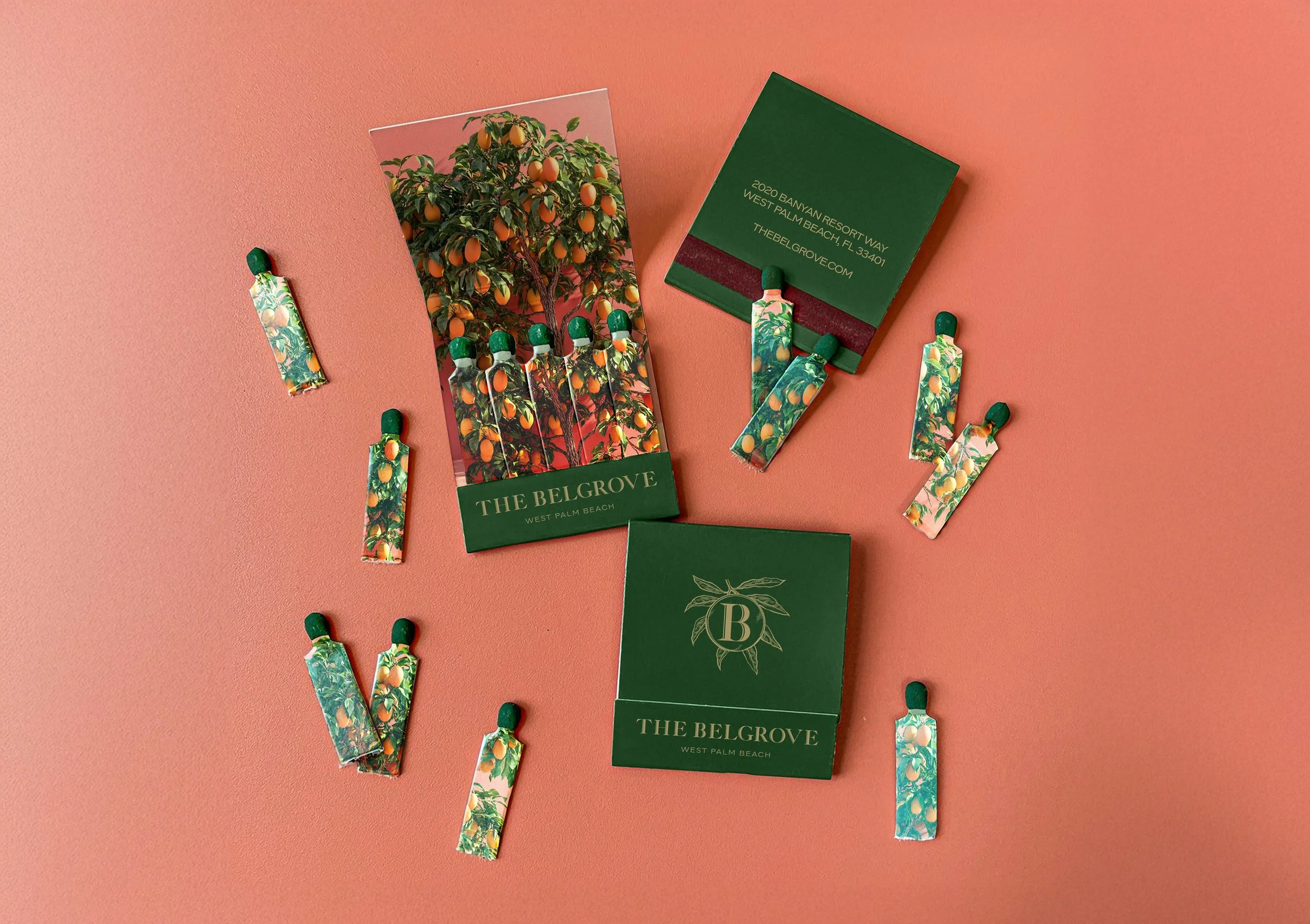A collection of packaging and sachets from The BelGrove, West Palm Beach, featuring a citrus-themed design with oranges and leaves. Includes two boxes with green and gold lettering, one with an image of an orange tree, and several individual sachets with matching design, all arranged on a pink surface.