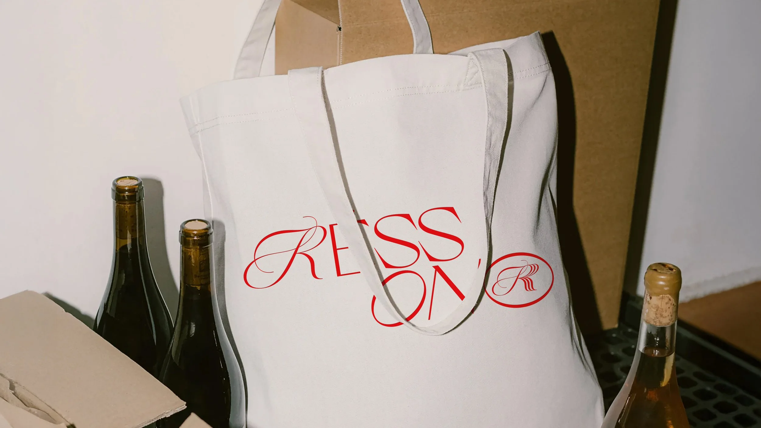 A white tote bag with red text and logo resting against a cardboard box, with three bottles of beer nearby.