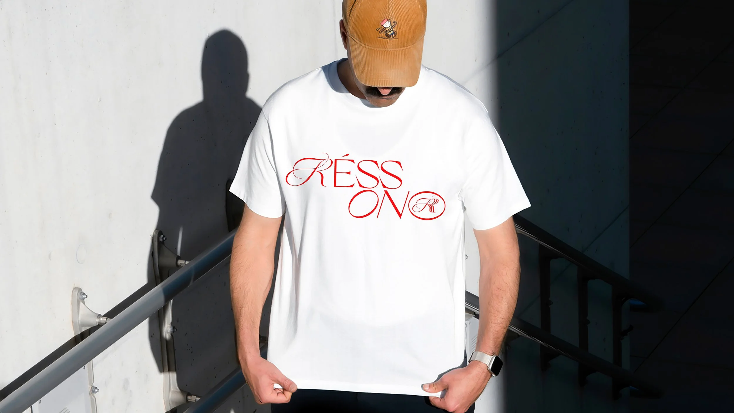 Person wearing a beige cap with character illustration, white T-shirt with red script text, against white and dark background.