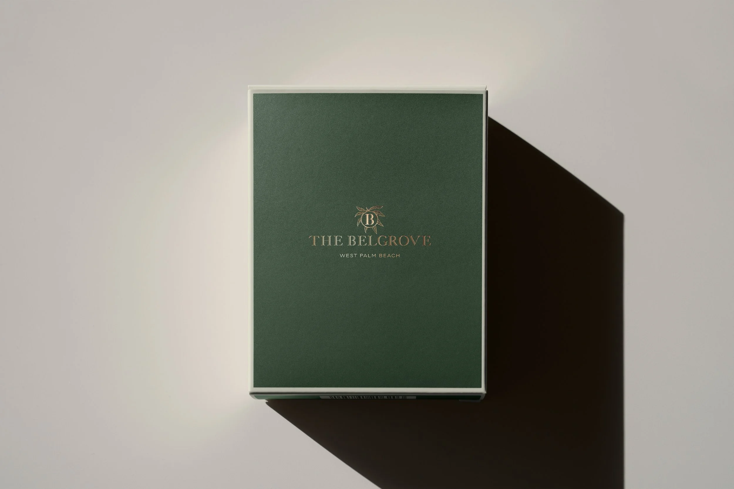 Green box with gold and red text reading 'The BelGrove, West Palm Beach' and a monogram 'B' with a leaf design in the center, casting a shadow on a white background.
