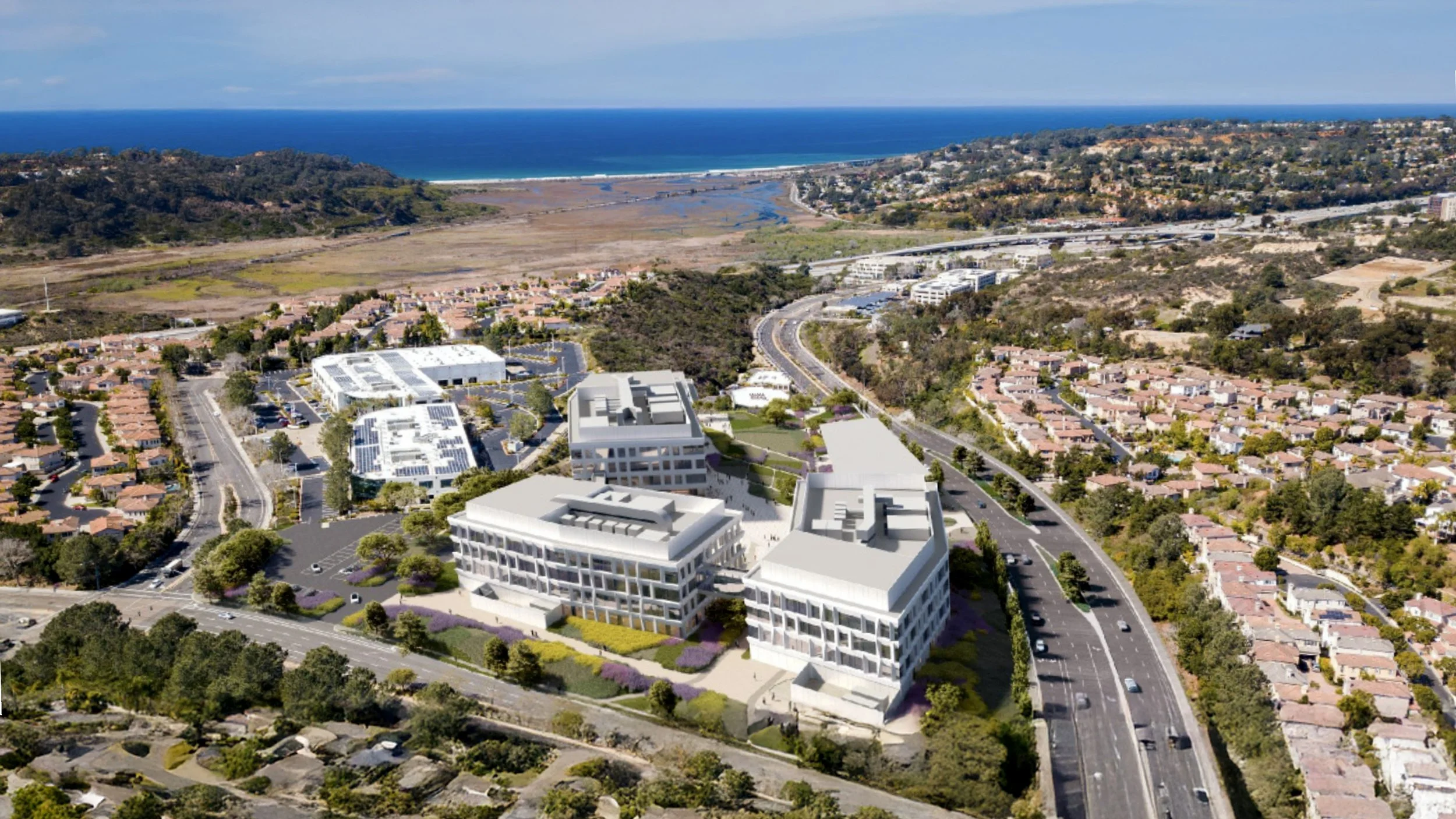 Aerial view of a coastal neighborhood with modern white buildings, residential houses, and a highway, with the ocean and hills in the background.