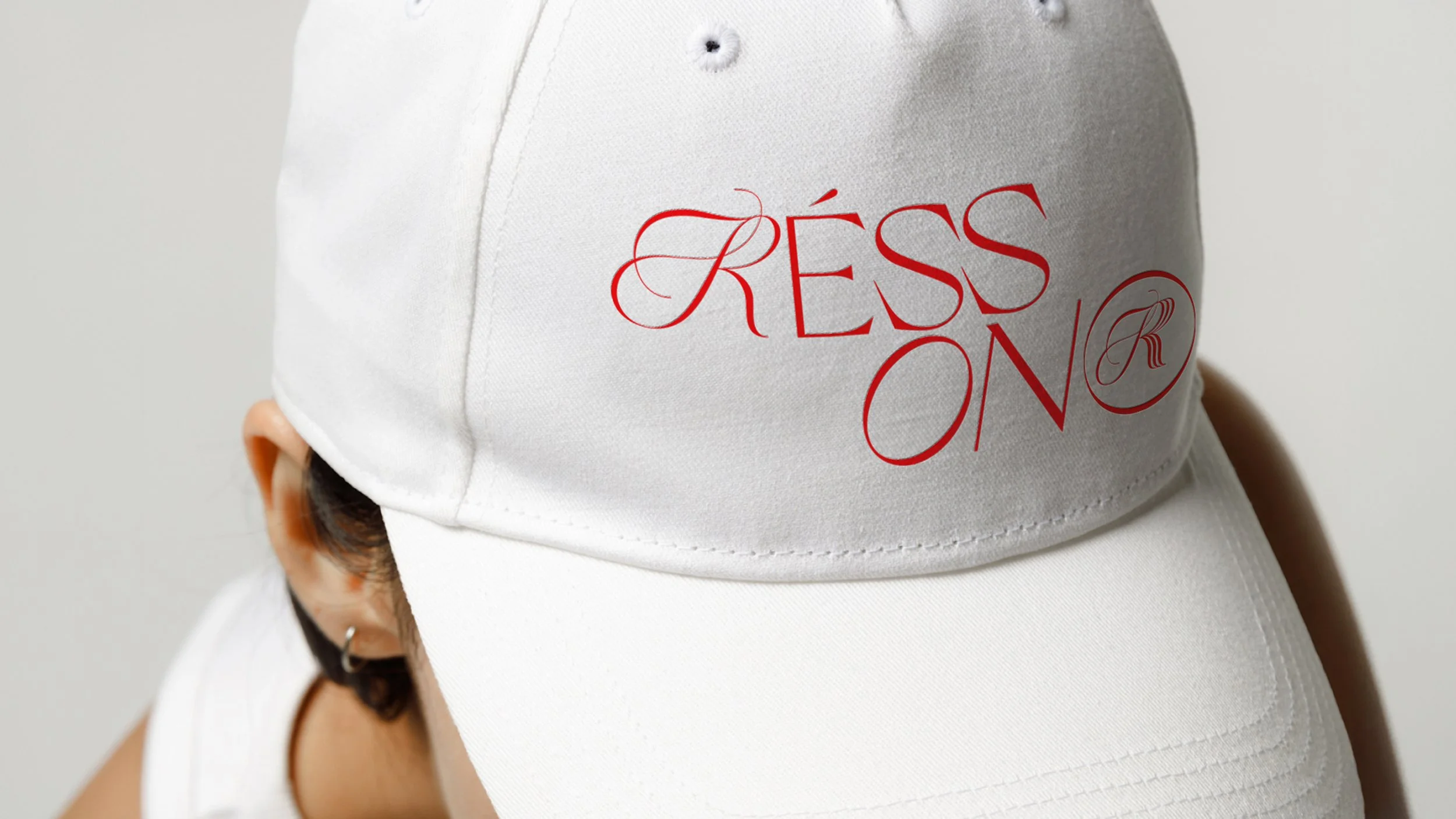 White cap with red embroidered text that reads 'FASHION' with a stylistic letter 'R' inside a circle on front, worn by a person with short dark hair and a hoop earring.