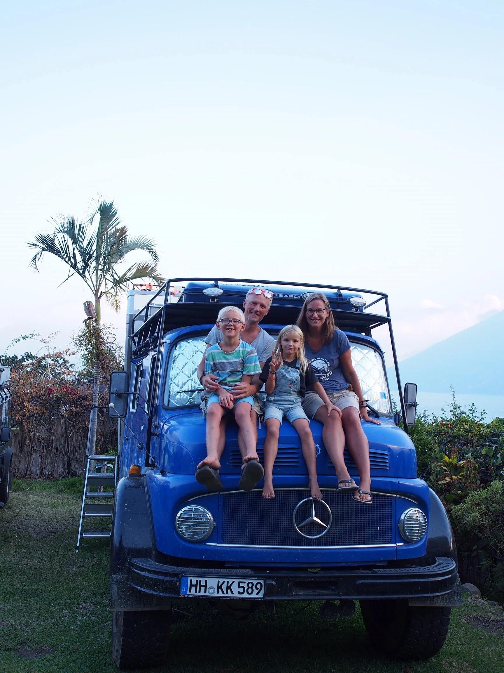 Familie sitzt auf der Motorhaube eines blauen Mercedes-Benz G-Wagen, im Hintergrund sind Palmen, Bäume und Berge zu sehen.