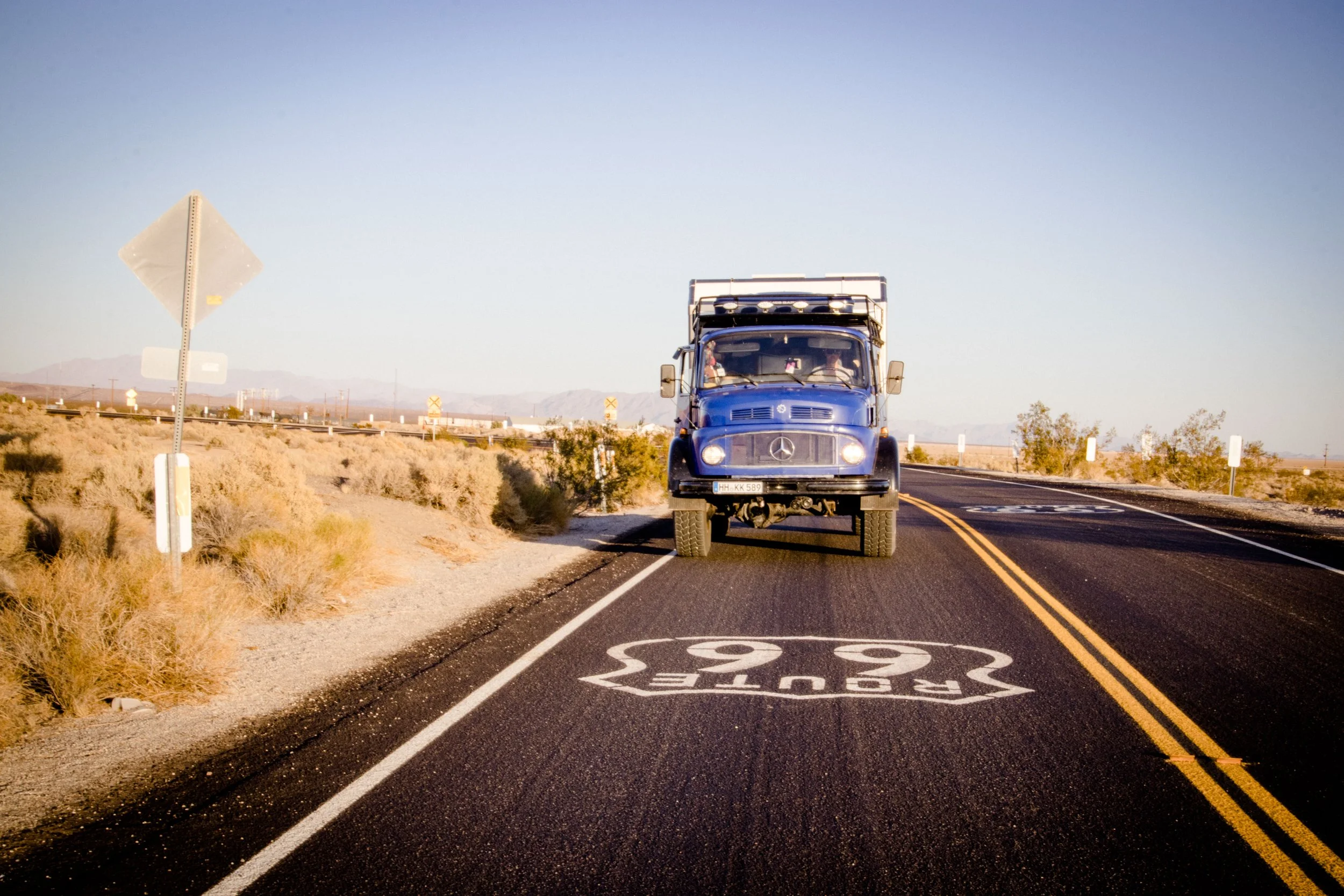 Ein blauer Mercedes-Benz LKW fährt auf einer zweispurigen Straße durch eine Wüstenlandschaft mit Bergen im Hintergrund. Auf der Straße ist ein Schild mit dem Schriftzug 'Route 66' sichtbar.