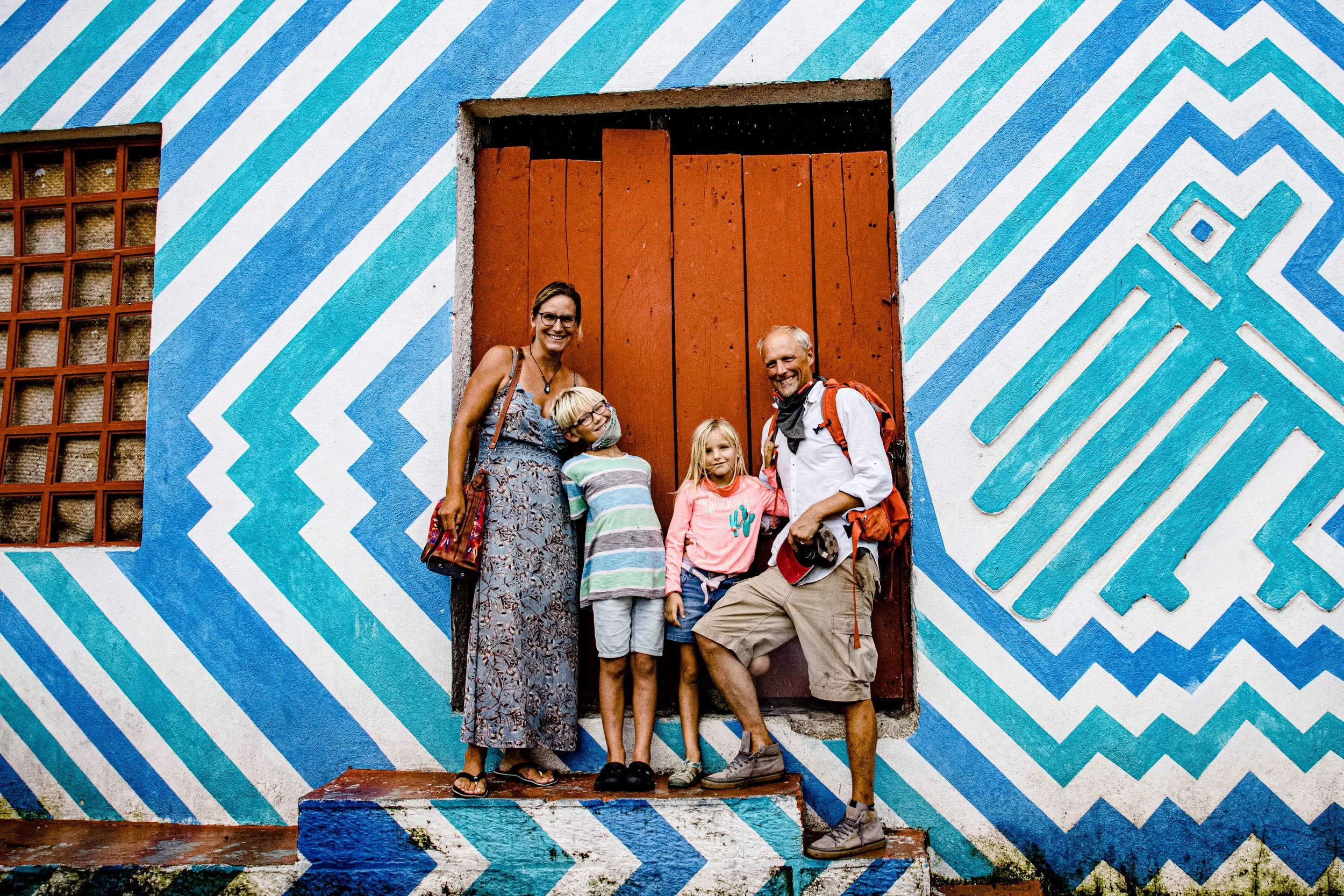 Eine Familie steht vor einer bunten mit blauen und weißen Mustern bemalten Wand und einem roten Holzfensterladen.