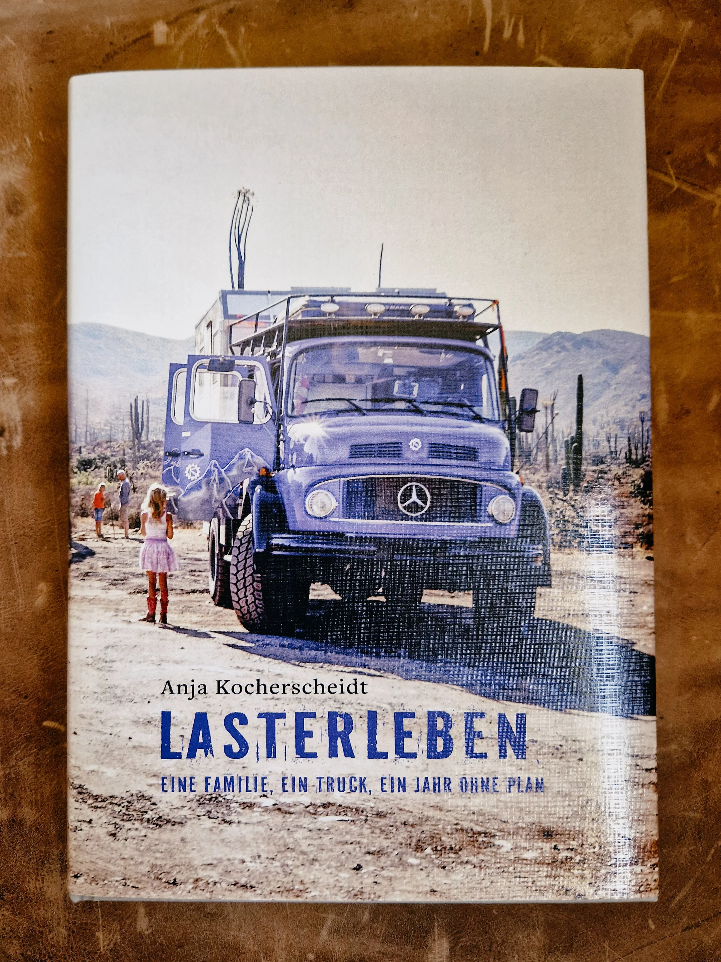 Buchdeckel mit einem Foto eines Lastwagens in einer Wüstenlandschaft in Baja California, im Vordergrund sind Kinder zu sehen. Der Titel des Buches lautet 'LASTERLEBEN' und die Autorin ist Anja Kocherscheidt.