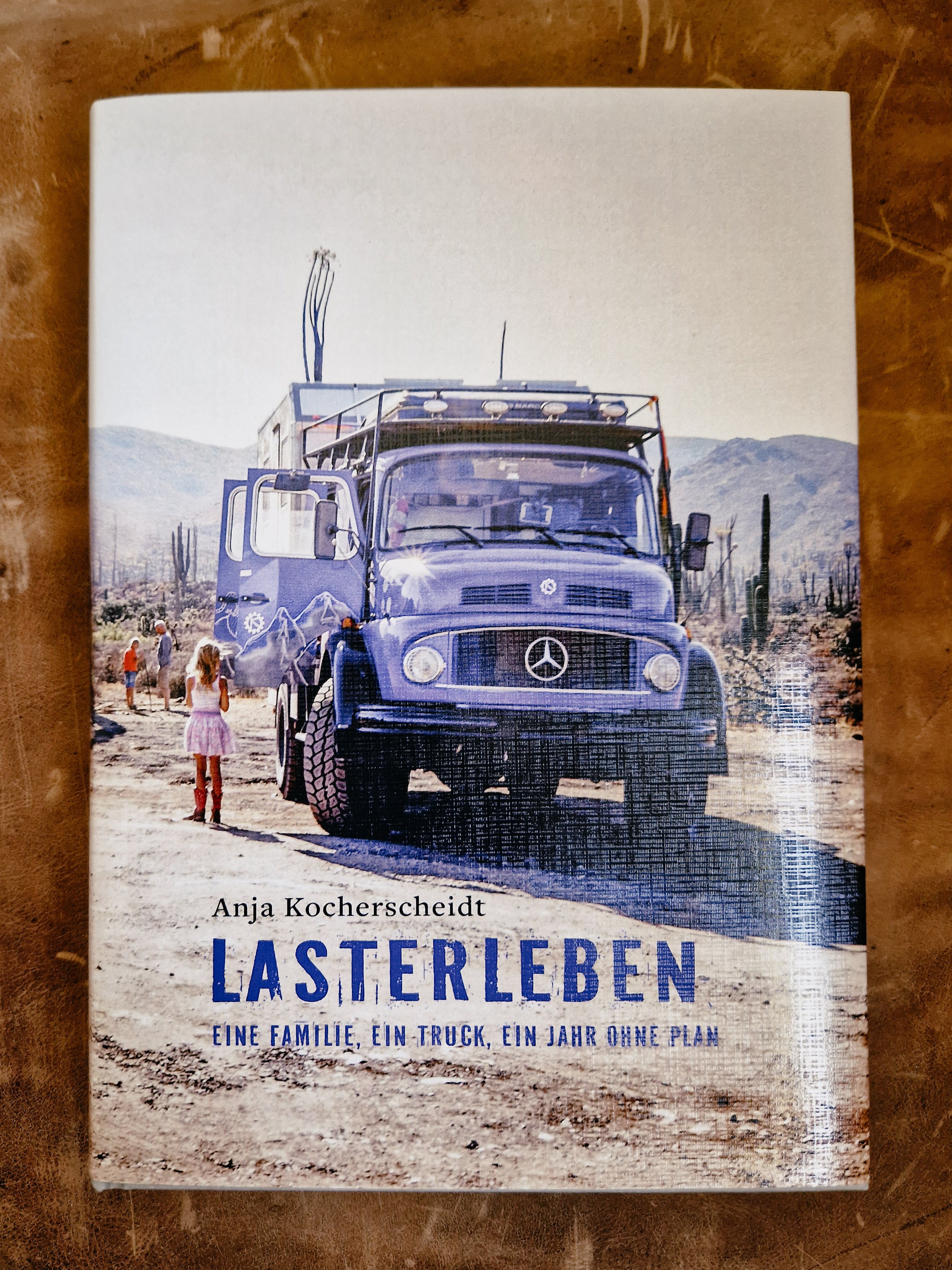 Buchcover von "Lasterleben" zeigt einen LKW in einer Wüstenlandschaft in Baja California mit Bergen im Hintergrund, darunter Kinder und Erwachsene, die den LKW betrachten.