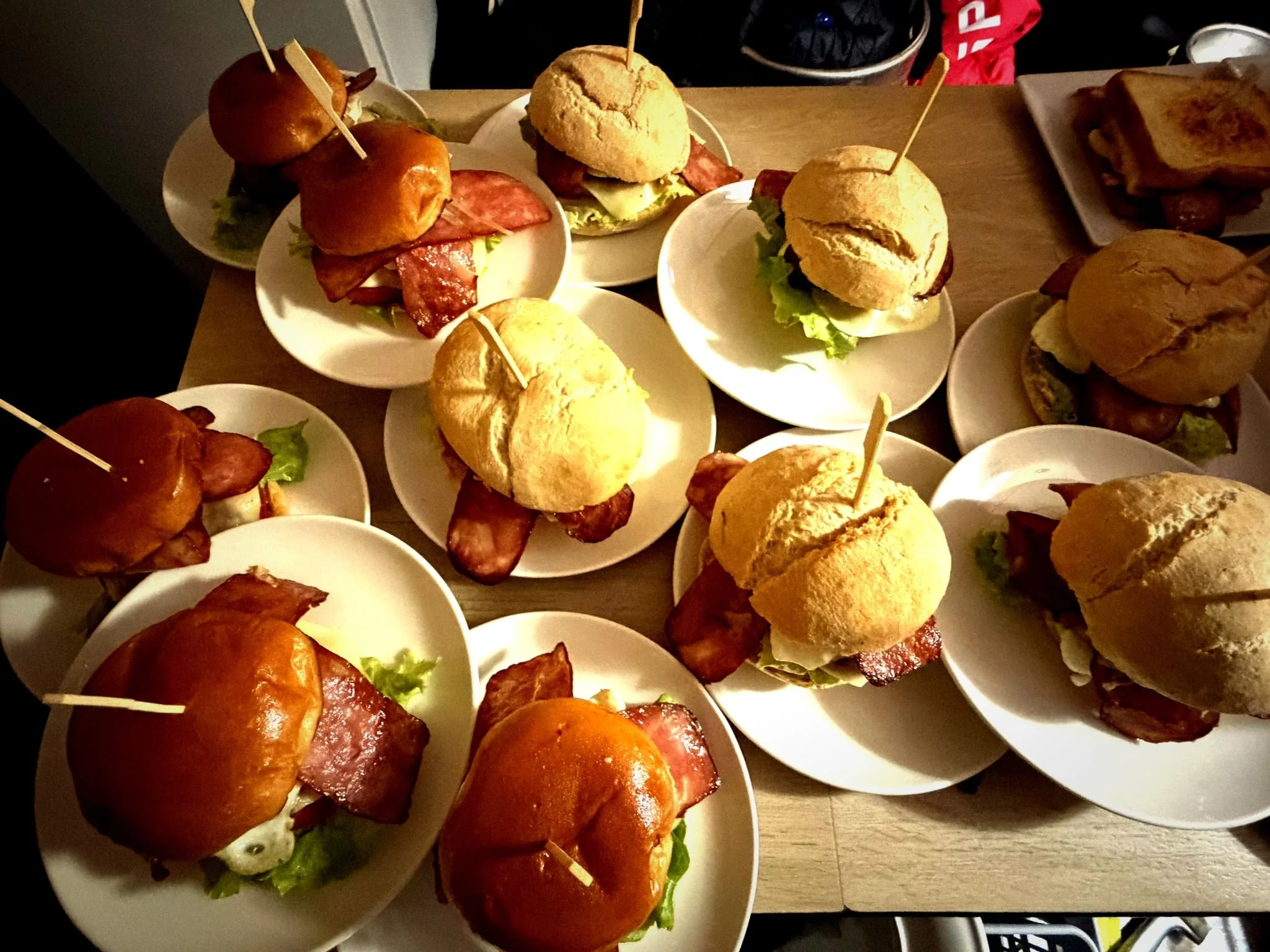 Varias mini hamburguesas con pan, carne, tocino, lechuga, y jitomate en platos blancos sobre una mesa de madera, algunas con palillos sujetando los ingredientes.