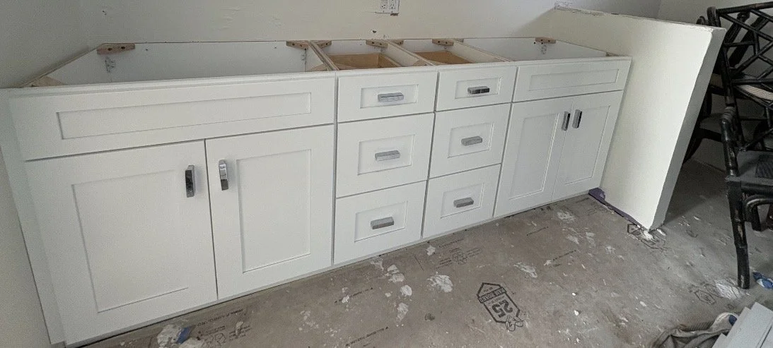 White kitchen cabinet with multiple drawers and doors, installed against a wall, with visible construction debris on the floor.