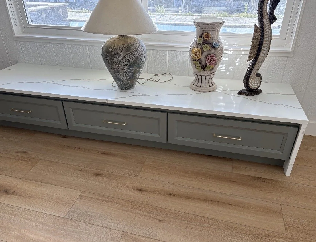 A white marble-top console table with three painted drawers, placed in front of a window. On the table, there is a ceramic table lamp with a beige shade, a decorative floral vase, and a seahorse sculpture. The floor is wooden, and the window has whit