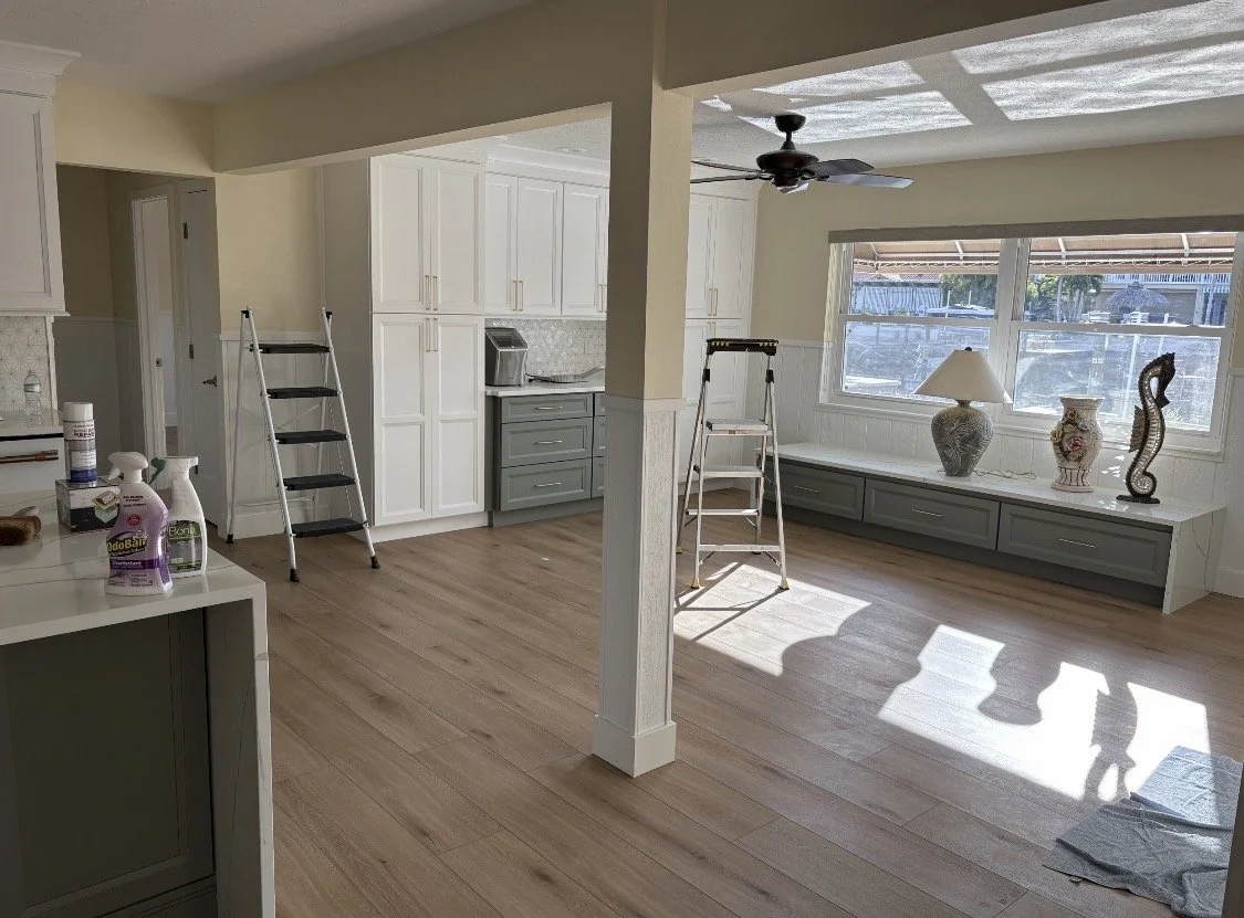 Empty living room and kitchen area with light wood flooring, white cabinetry, and decorative vases on built-in window bench. Two stepladders are in the room, and sunlight is streaming through the window.