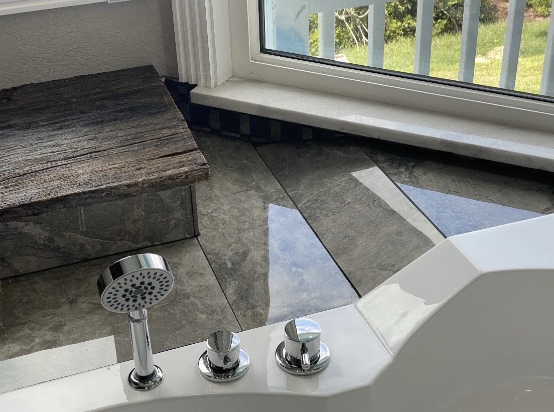 Close-up of a bathroom shower area with a white bathtub, two chrome knobs, and a handheld showerhead. The floor tiles are gray marble, and there is a large window with a view of greenery outside.