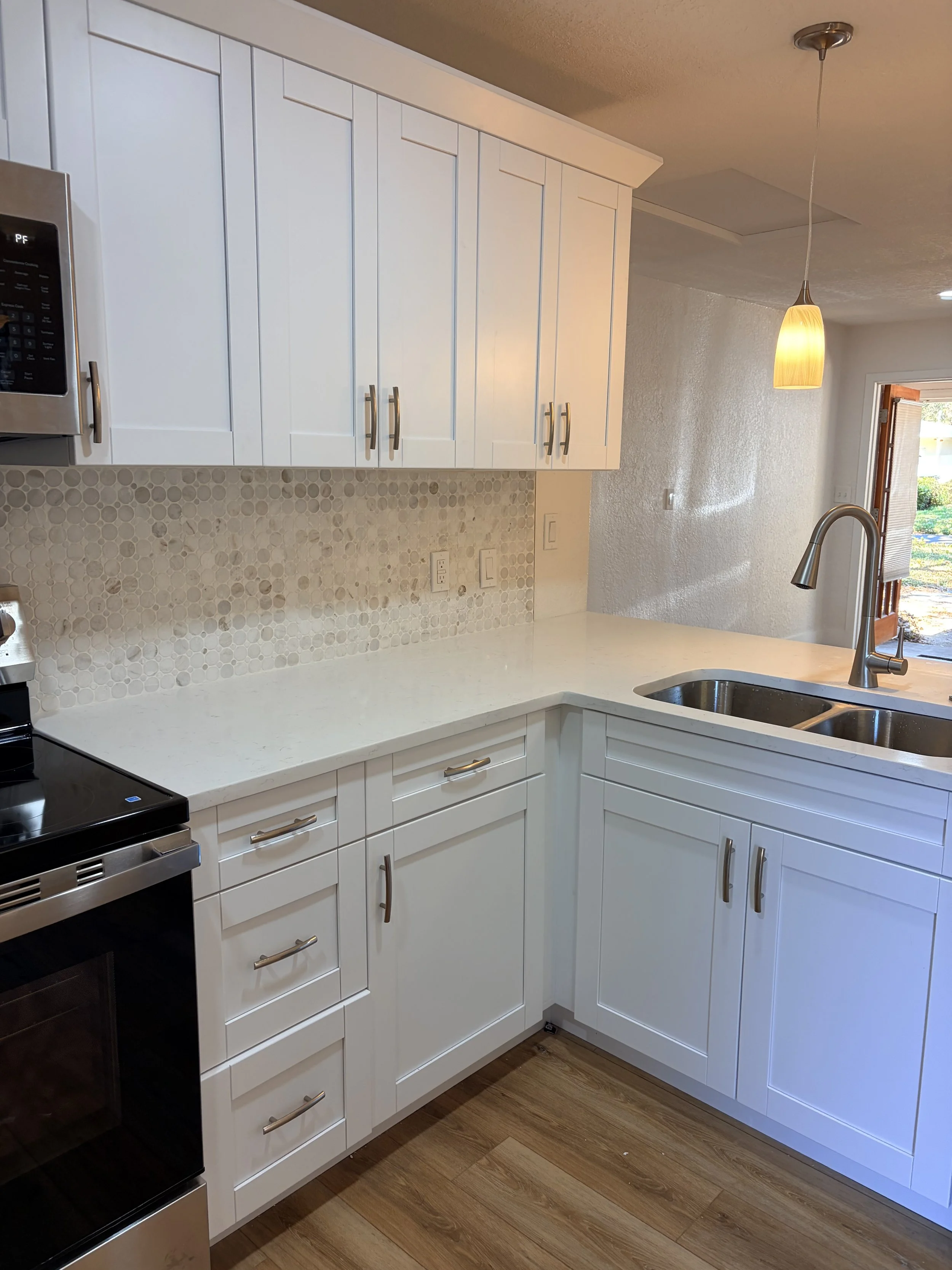 Modern kitchen with white cabinets, a light-colored countertop, a backsplash with circular tile pattern, a stainless steel microwave, and a stainless steel sink with a curved faucet. There is also a pendant light hanging from the ceiling and a view of the door to the outside.