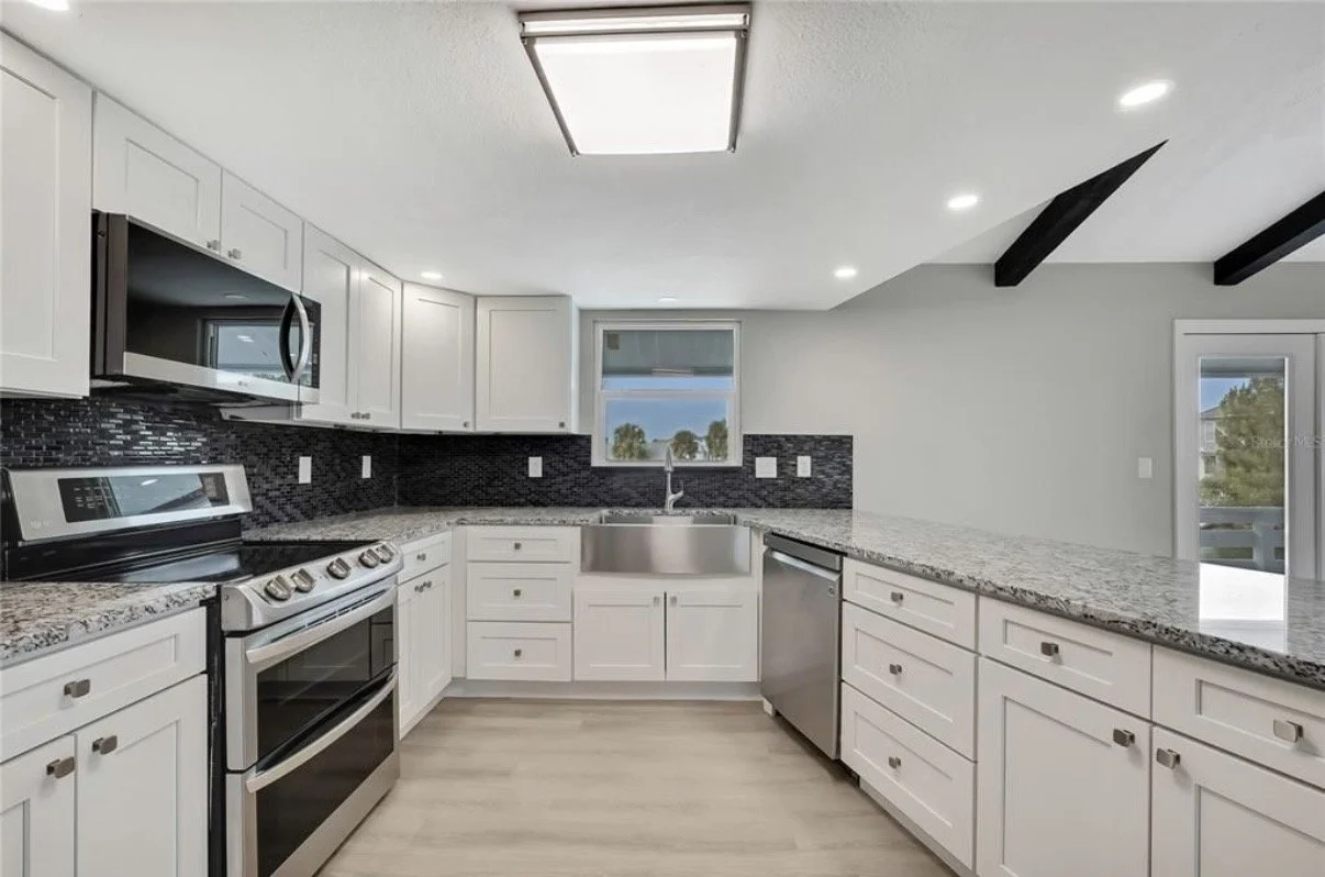 Modern kitchen with white cabinets, granite countertops, black backsplash, stainless steel appliances, and a small window above the sink, with a sliding door leading outside.