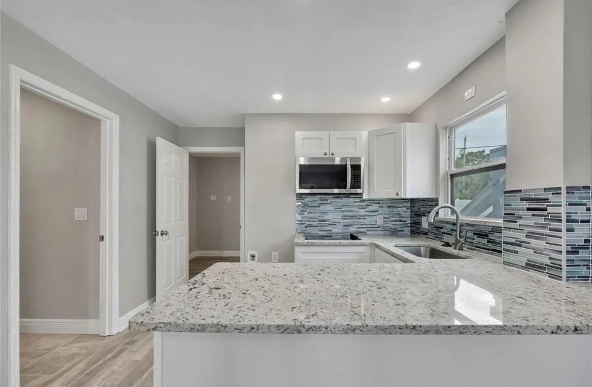 Modern kitchen with white cabinets, a granite countertop, a stainless steel microwave, and a window with a view outside. The wall behind the counter has a glass tile backsplash.