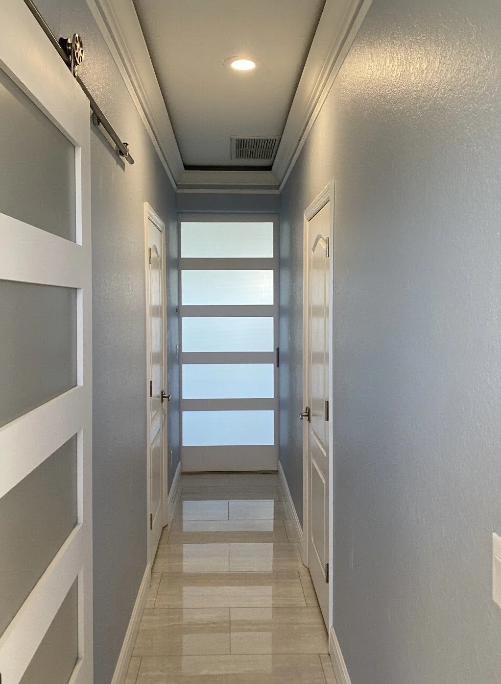 A hallway with light blue walls, three white doors, a frosted glass door at the end, and a skylight in the ceiling.