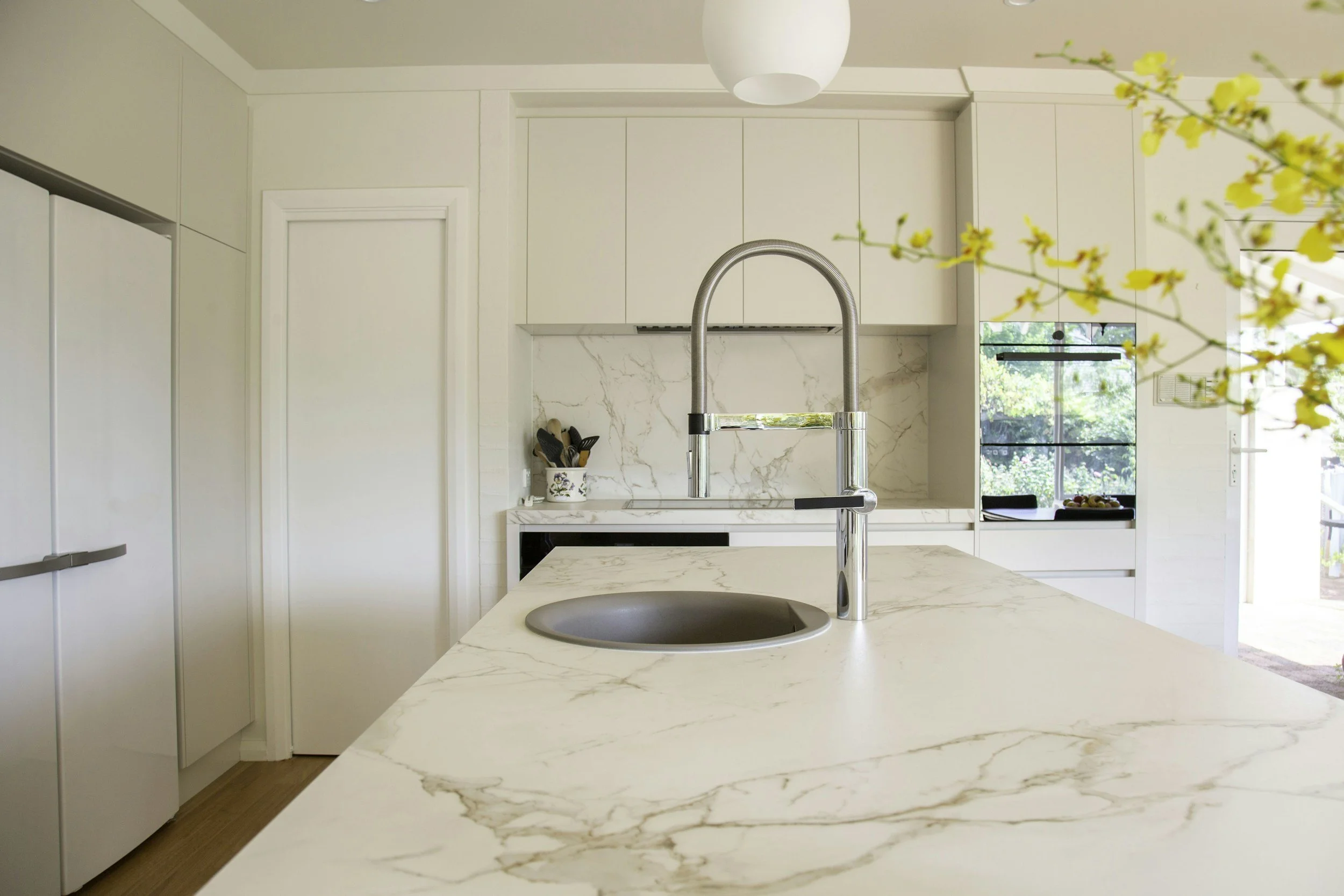 Modern kitchen with white marble countertops, a round sink, and a sleek, tall faucet. White cabinets and a marble backsplash are visible, with sunlight coming through a large window. Yellow flowers are slightly blurred in the foreground.