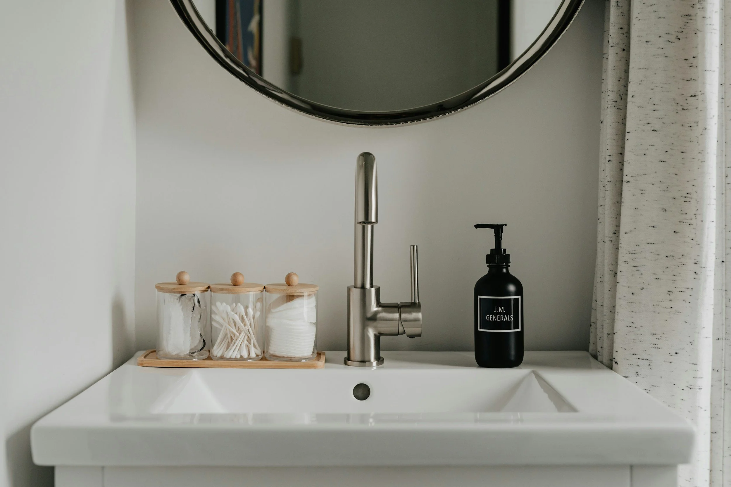 Minimalist kitchen sink area with a modern stainless steel faucet, a black soap dispenser labeled "J.M. Generals," and clear jars with wooden lids containing cotton swabs, cotton pads, and paper straws, set against a white countertop and light-colored tiled backsplash.