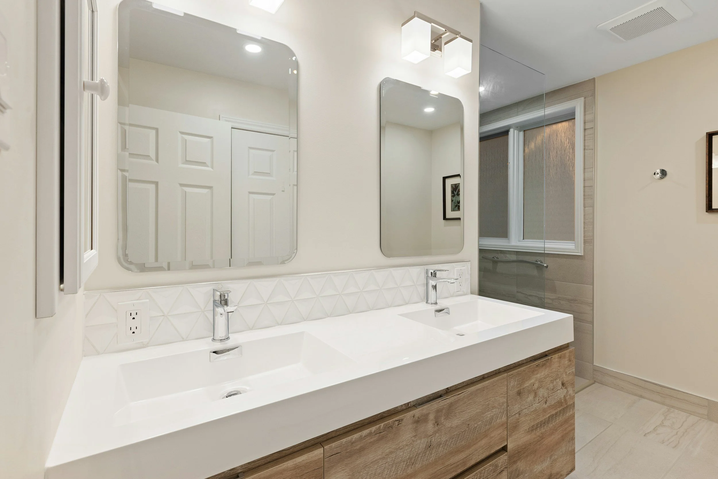 Bathroom vanity with dual sinks, large mirrors, modern light fixtures, and a window in the background.