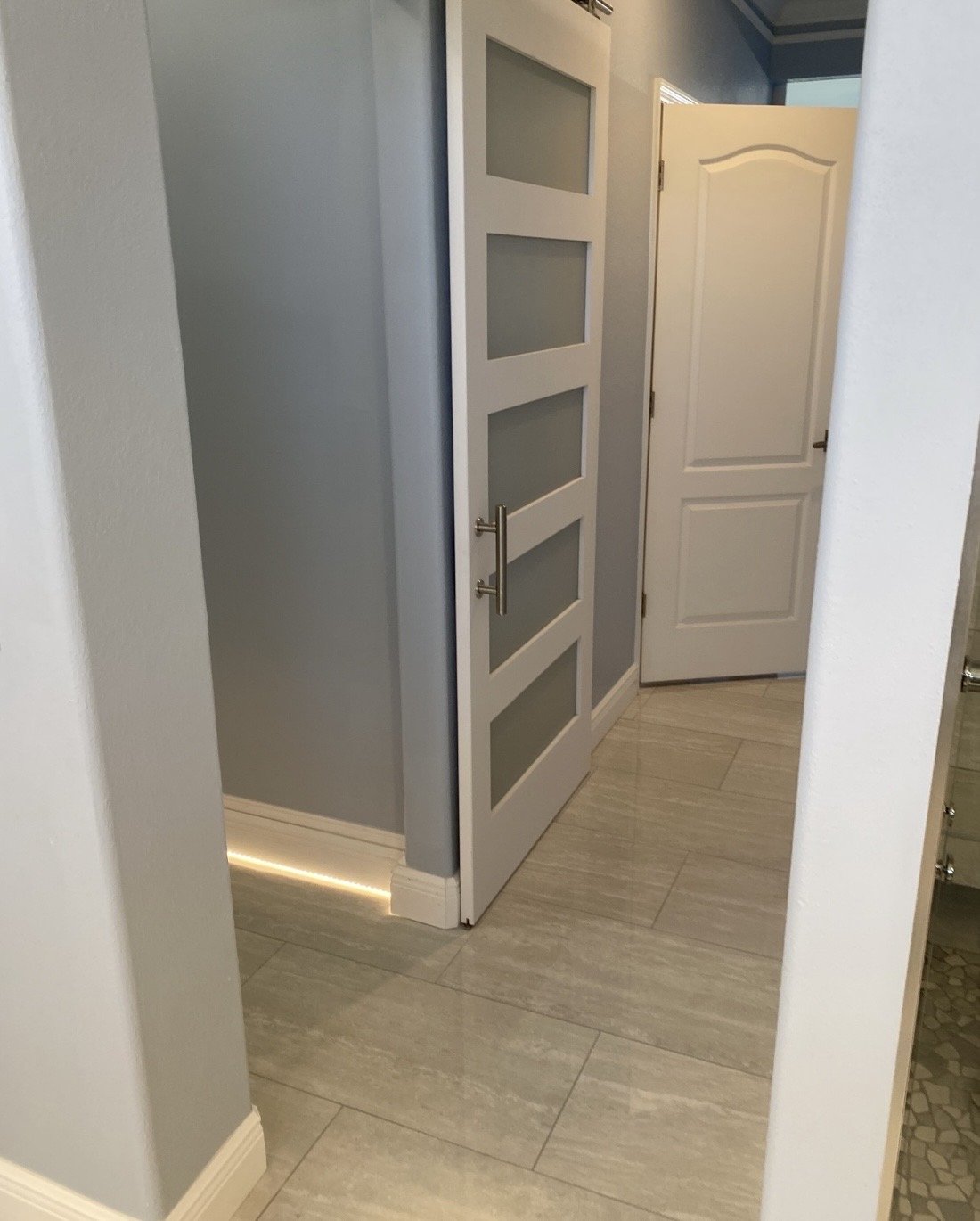 A hallway with light-colored tiles, gray walls, and two white doors, one with glass panels and one plain, with a small gap at the bottom and a strip of light visible underneath.