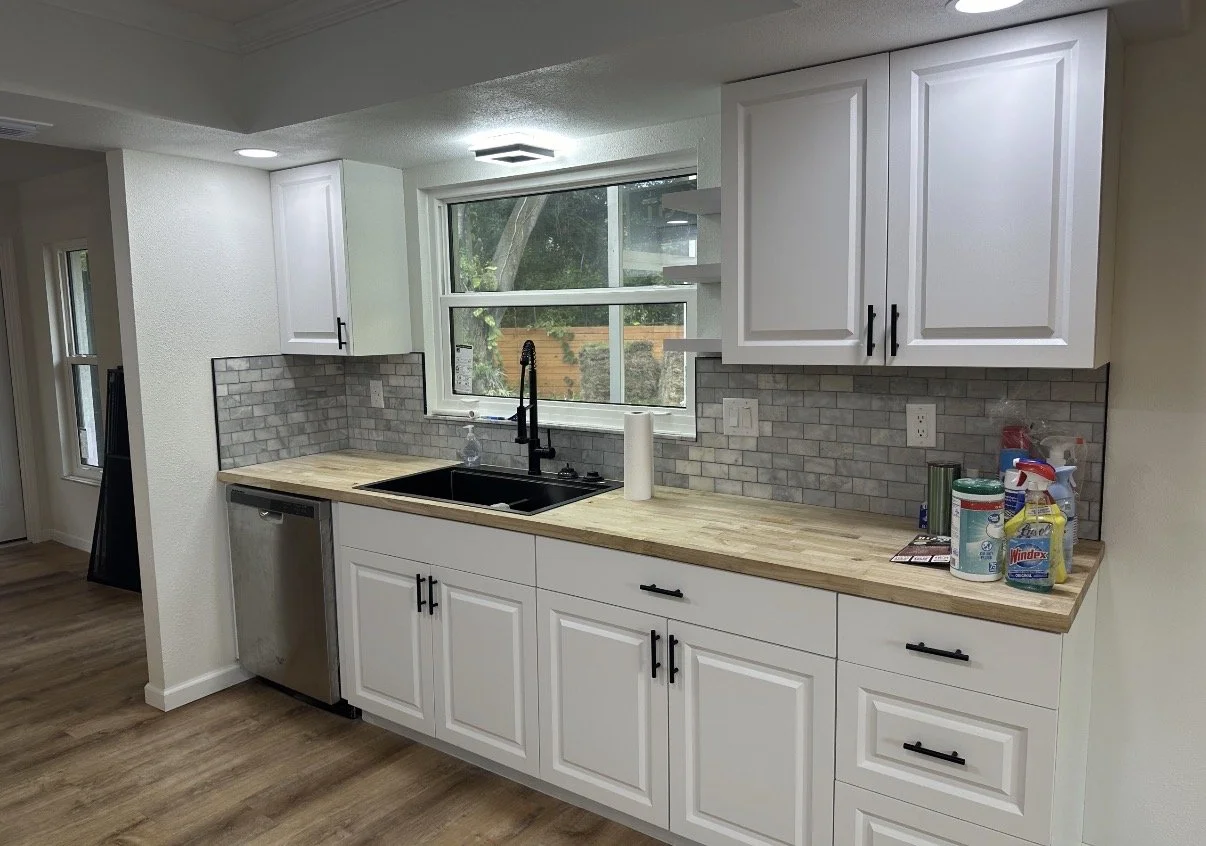 Kitchen with white cabinets, wooden countertop, gray subway tile backsplash, window above sink, black faucet, and various cleaning supplies on the counter.