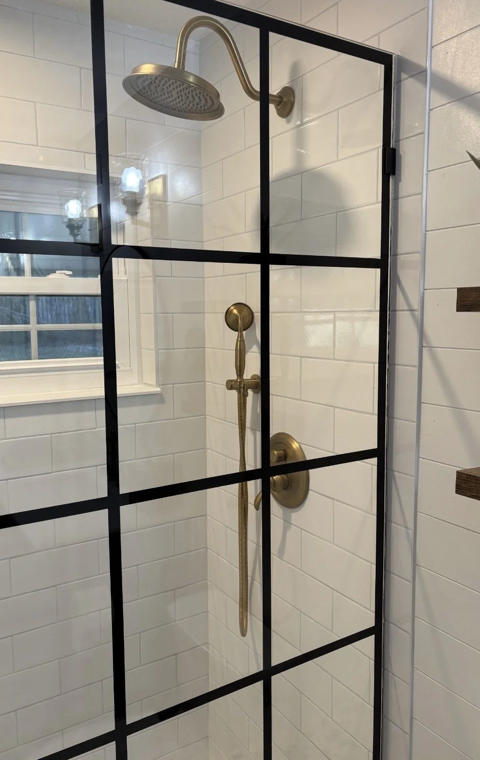 Shower with a brass showerhead and handheld spray inside a glass enclosure with black framing, white tiled walls, and a small window.