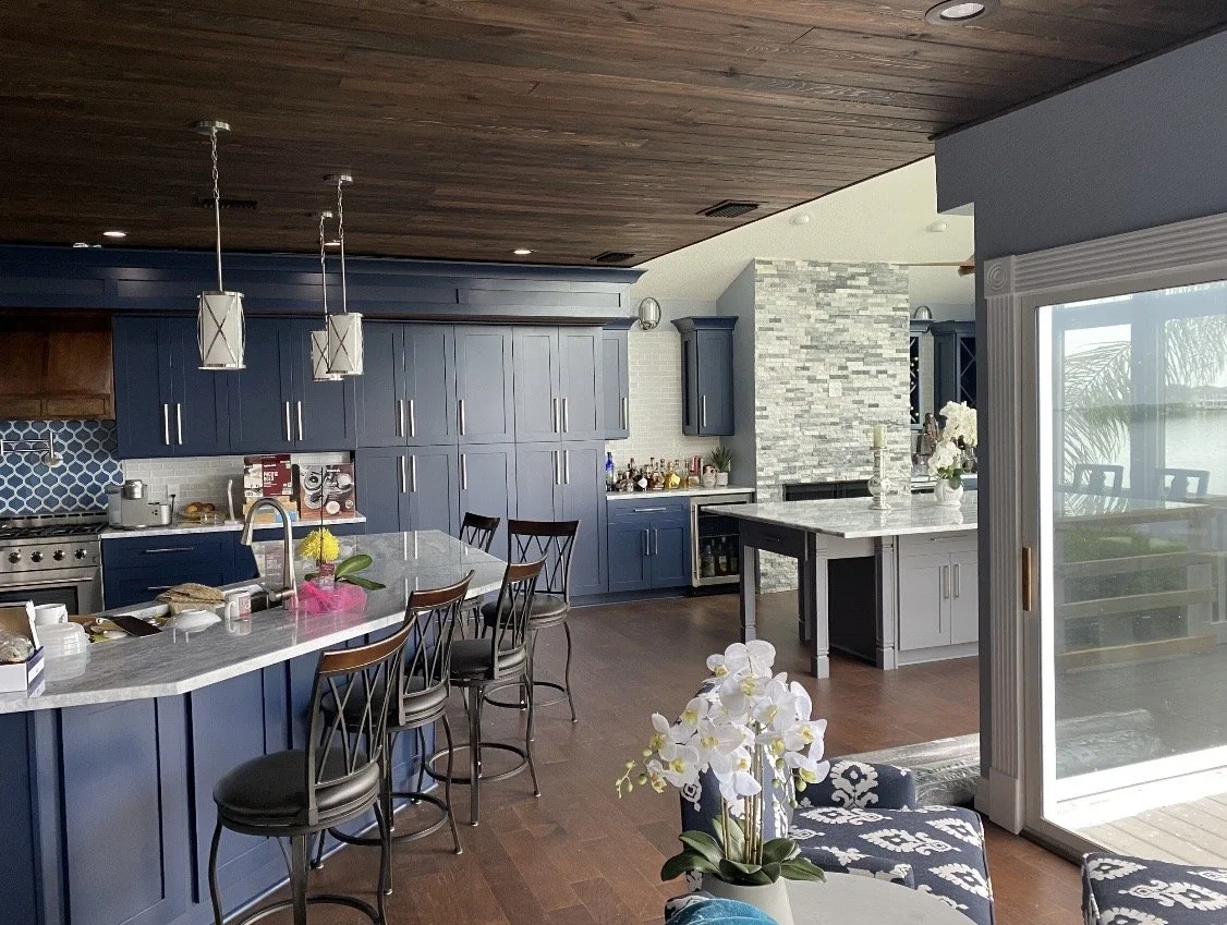 Modern kitchen with blue cabinets, gray island, and wooden ceiling. Bar chairs around island, sliding glass door to patio, and flowers on table.