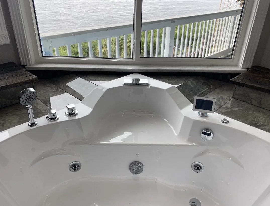 A luxurious white bathtub with multiple jets, located near a glass door with a balcony view of the ocean and greenery outside.