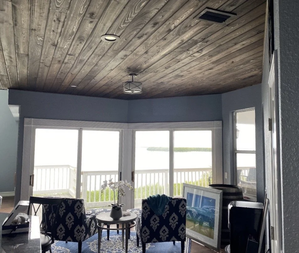 Living room with large windows overlooking water, wooden ceiling, modern light fixture, and seating area with patterned chairs and a small table.