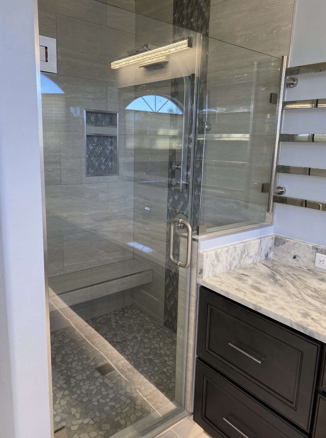 Bathroom with a glass-enclosed shower with a bench, pebble stone floor, wall niche, and a marble vanity countertop with dark cabinetry.