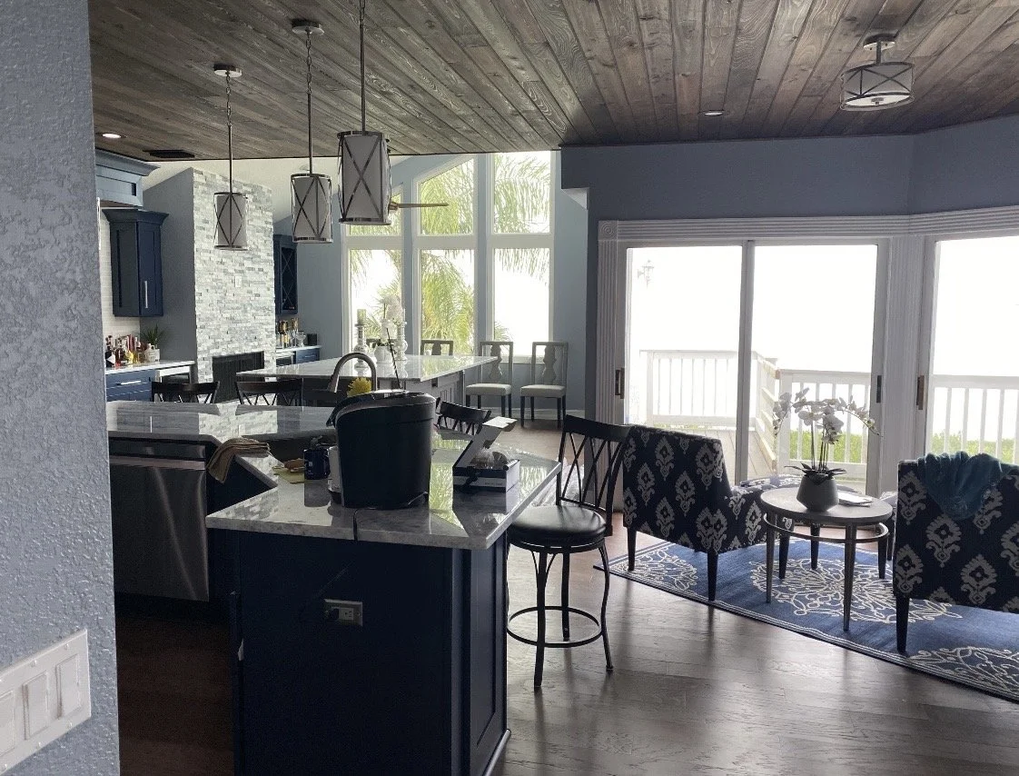 Open-concept living room and kitchen area with large windows, blue and white decor, bar stools, and outdoor balcony