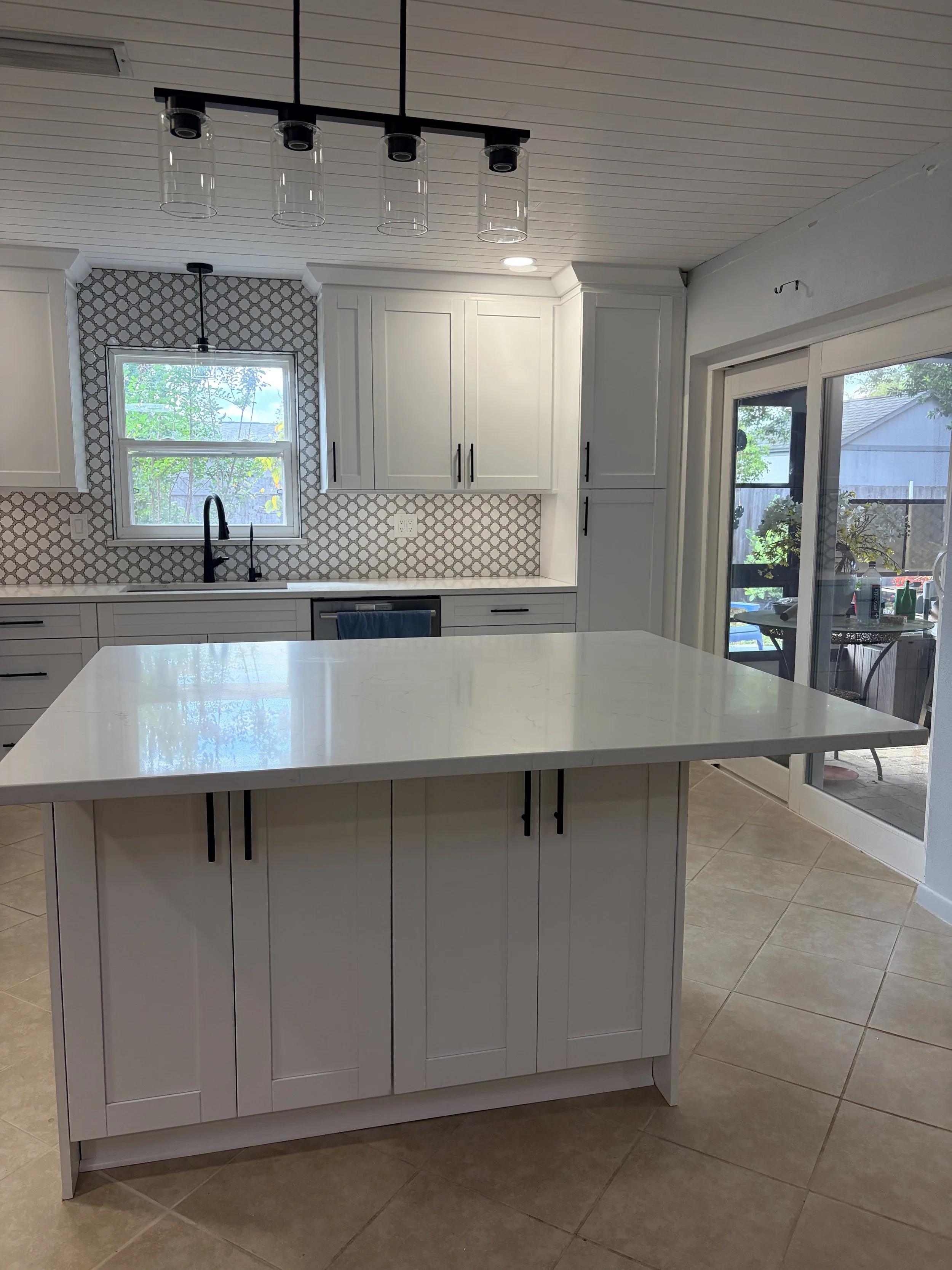 Modern kitchen with white cabinets, a kitchen island with a white countertop, tiled flooring, a window above the sink, sliding glass door leading outside, and pendant lighting fixtures.