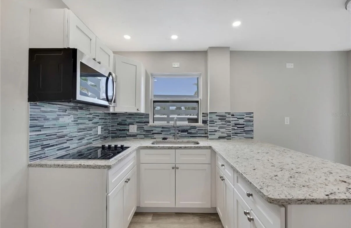 Modern kitchen with white cabinets, granite countertops, colorful tile backsplash, microwave, cooktop, and a window above the sink.