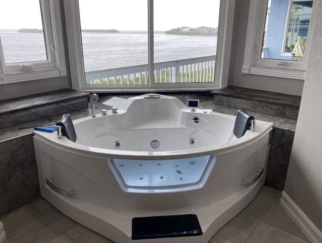 White corner jacuzzi with headrests and waterfalls, positioned in front of large glass windows overlooking a body of water with land in the distance, in a bright bathroom with tiled floors.