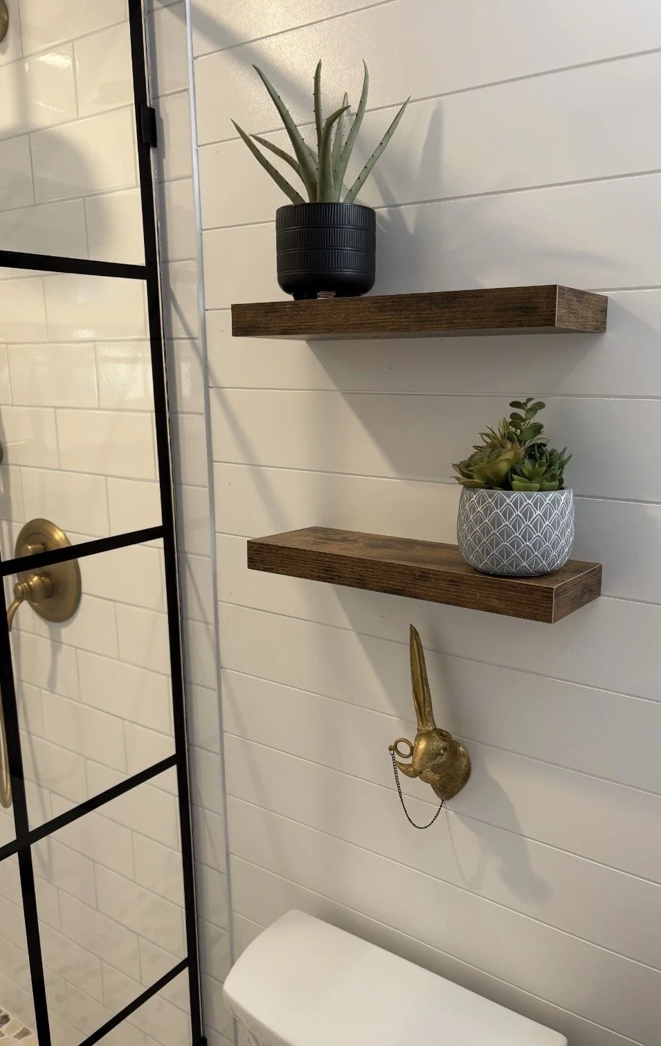 Decorative bathroom wall with two wooden shelves holding potted plants, golden rabbit wall ornament, and part of a black-framed glass shower door.