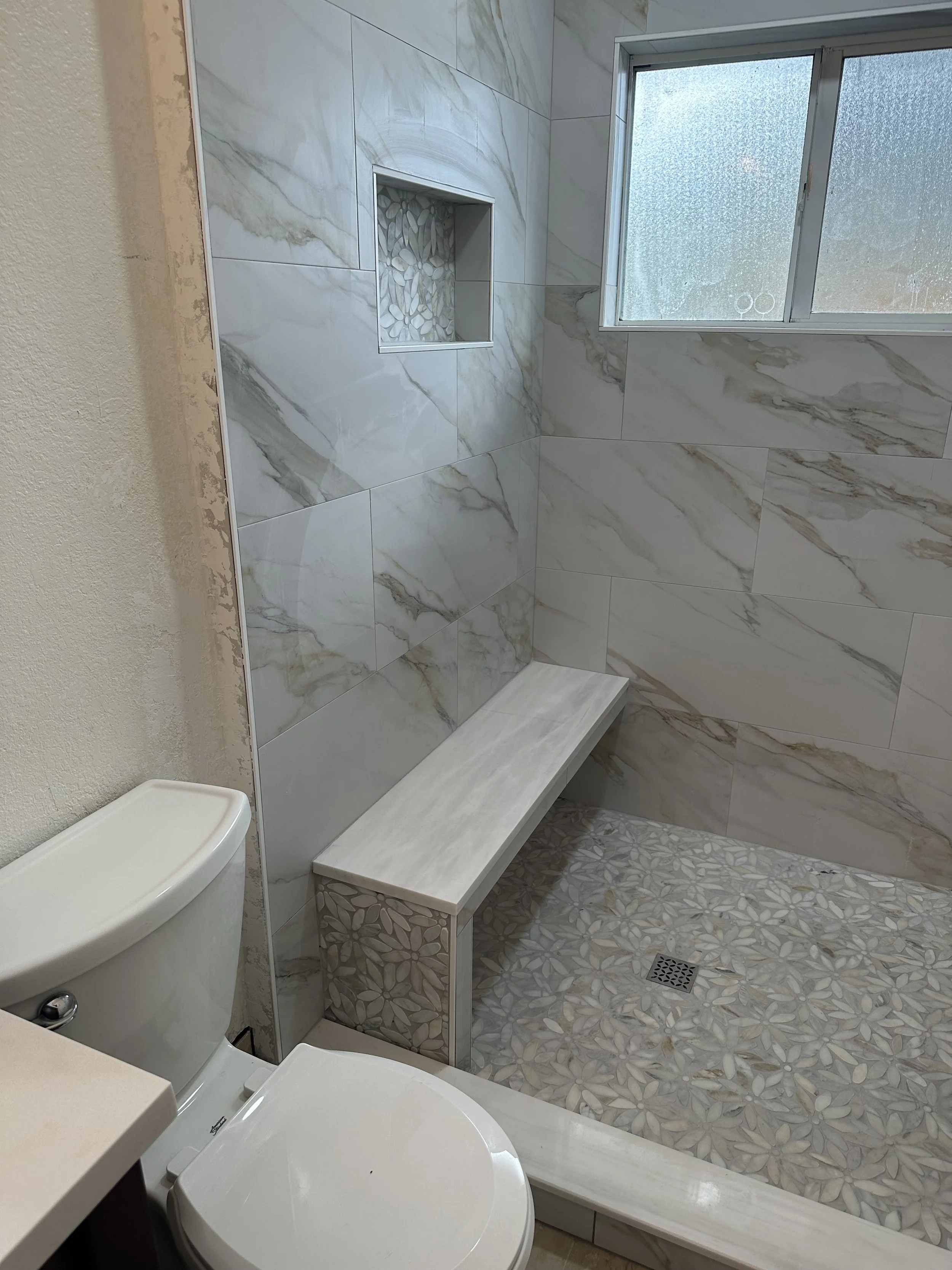 A bathroom with marble tile walls and pebble mosaic floors. A small shower area with a built-in niche with pebble stones, a window with frosted glass, a bench with shell mosaic pattern, and part of a toilet are visible.