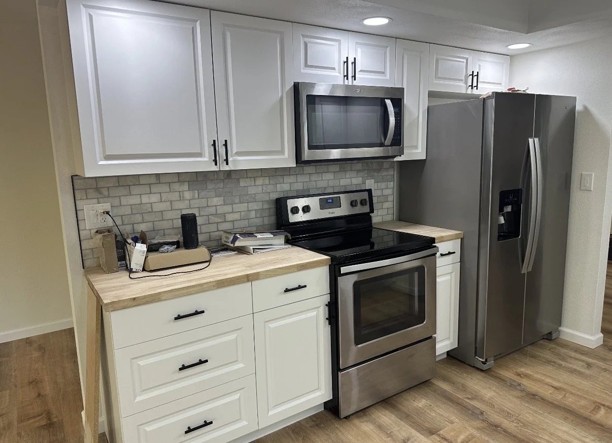 Kitchen with white cabinets, stainless steel microwave, oven, and refrigerator, beige tile backsplash, wooden countertops, and wood flooring.