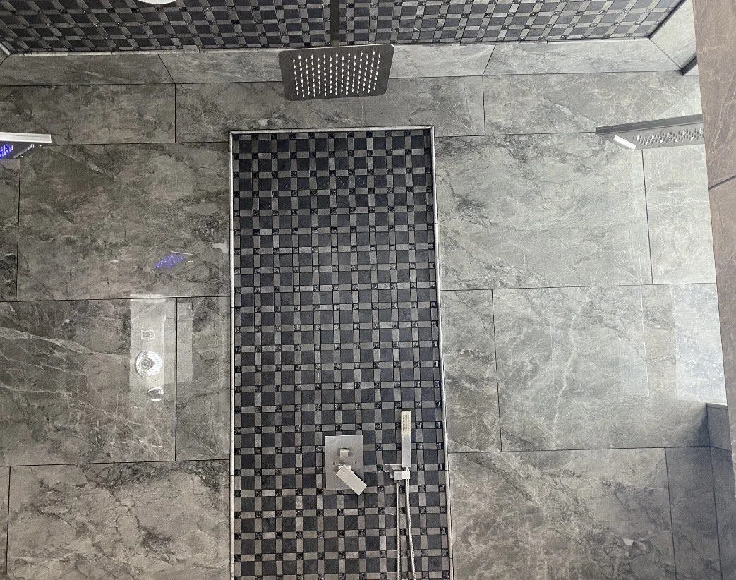 Two rain shower heads mounted on the ceiling, with a tiled floor featuring a central black and gray checkered pattern, and a metallic shower control panel with a hose and handheld shower.