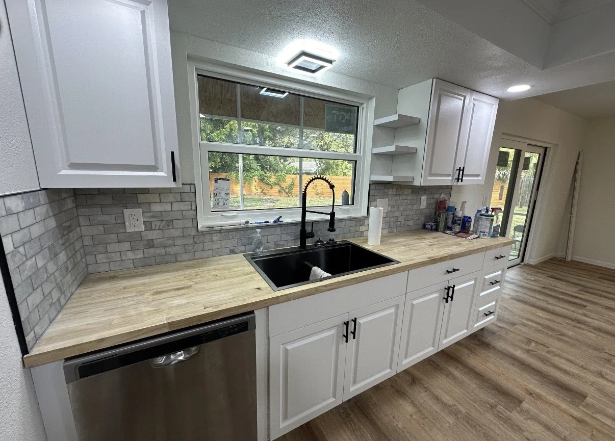 Kitchen with white cabinets, a black sink with a high-arc faucet, a wooden countertop, and a brick backsplash, featuring a window overlooking a backyard with trees and a sliding glass door leading outside.