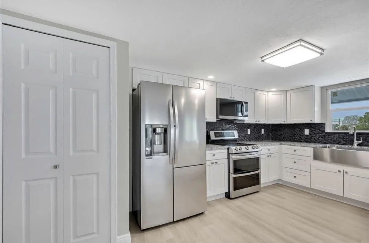 Kitchen with white cabinets, stainless steel refrigerator, microwave, oven, and sink, with black tile backsplash and light wood flooring.