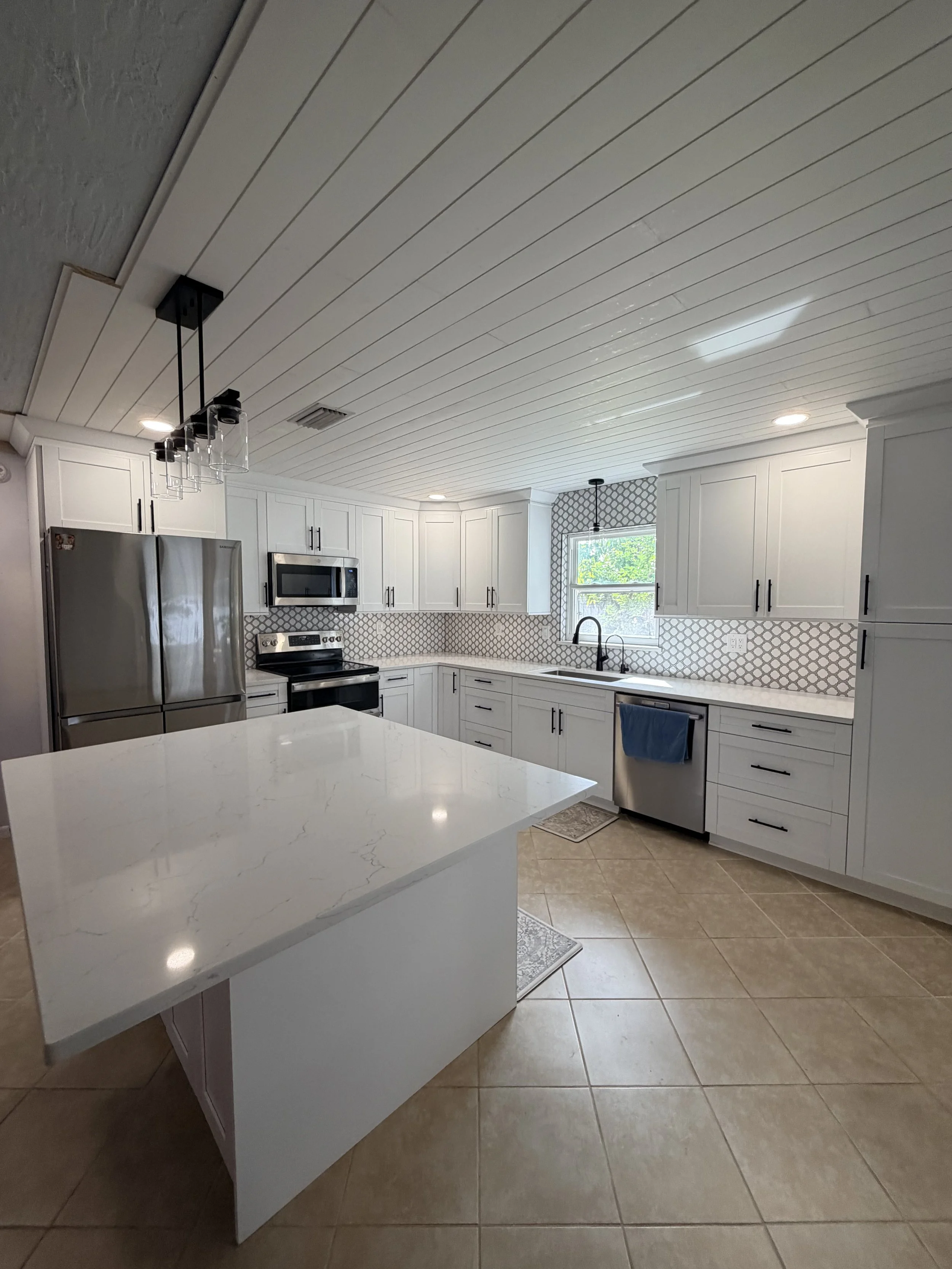 Beautiful modern kitchen with white cabinets, stainless steel appliances, and a white marble island. There are tiled floors and a window with a view outside.
