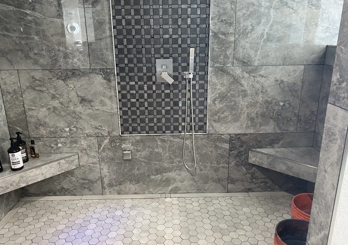 View of a modern shower with gray marble walls, a built-in corner ledge, a wall-mounted shower head, a handheld shower, and a mosaic tile accent wall. There are bottles of soap and shampoo on a marble shelf to the left, and red buckets on the floor t