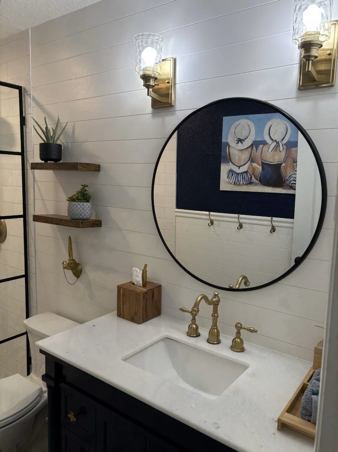 Bathroom with white paneled walls, two wall-mounted gold and glass light fixtures, a round mirror above the sink, a framed picture of two women in swimsuits with large sun hats, two wooden shelves with potted plants, a tissue box on the sink, and a s
