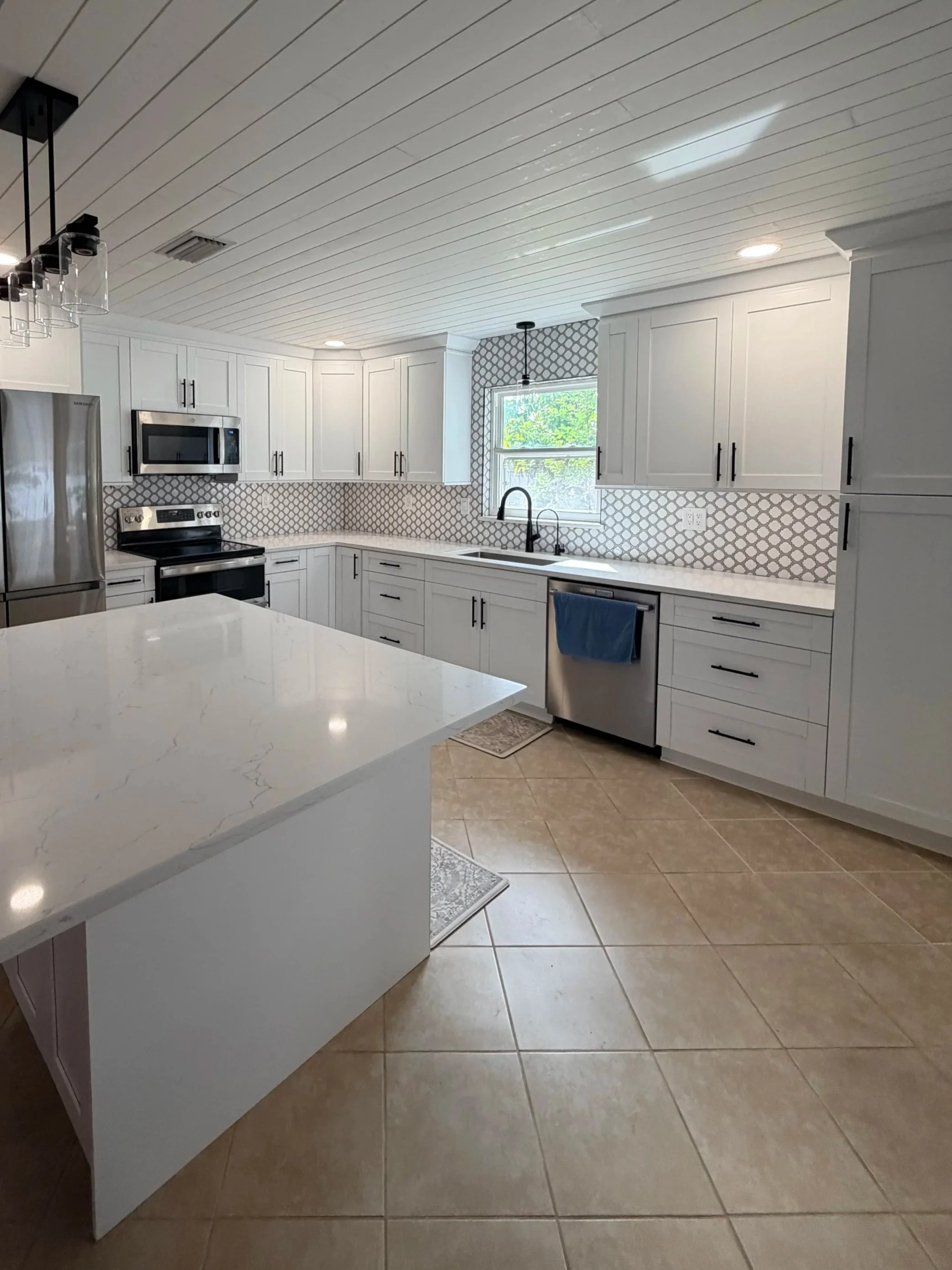 Modern kitchen with white cabinets, a white marble countertop island, stainless steel appliances, tile flooring, and a window with greenery outside.