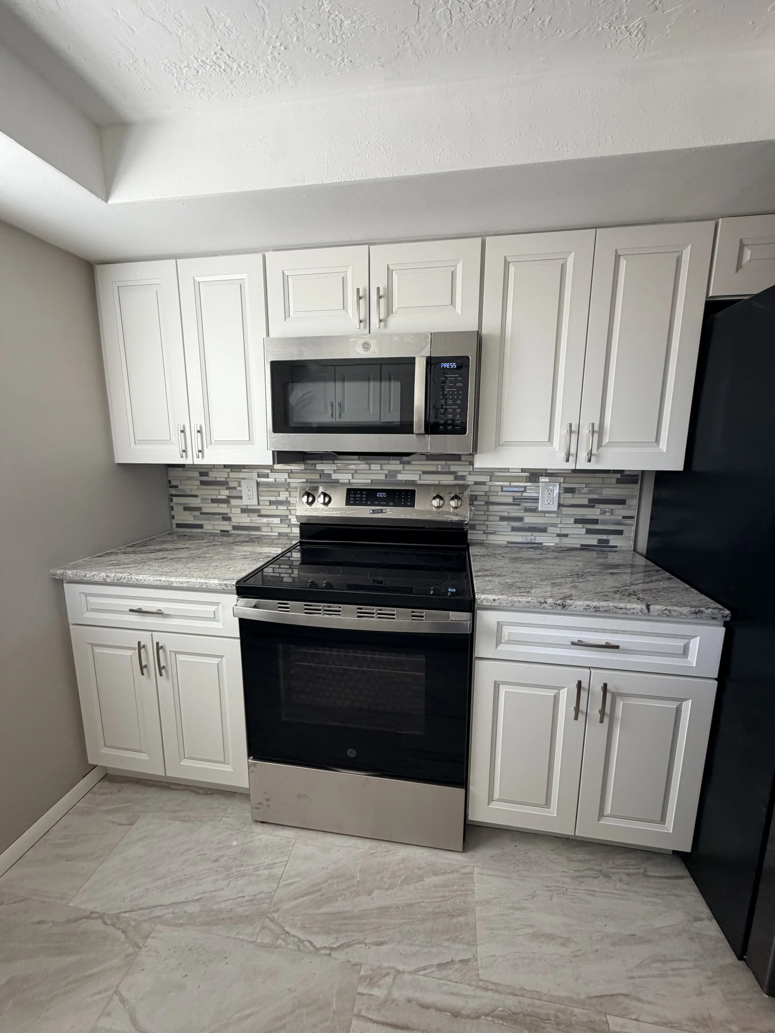A modern kitchen with white cabinets, a stainless steel microwave above a stove, granite countertops, and a decorative backsplash made of small rectangular tiles in gray, white, and black tones. The floor is light-colored with large tiles.