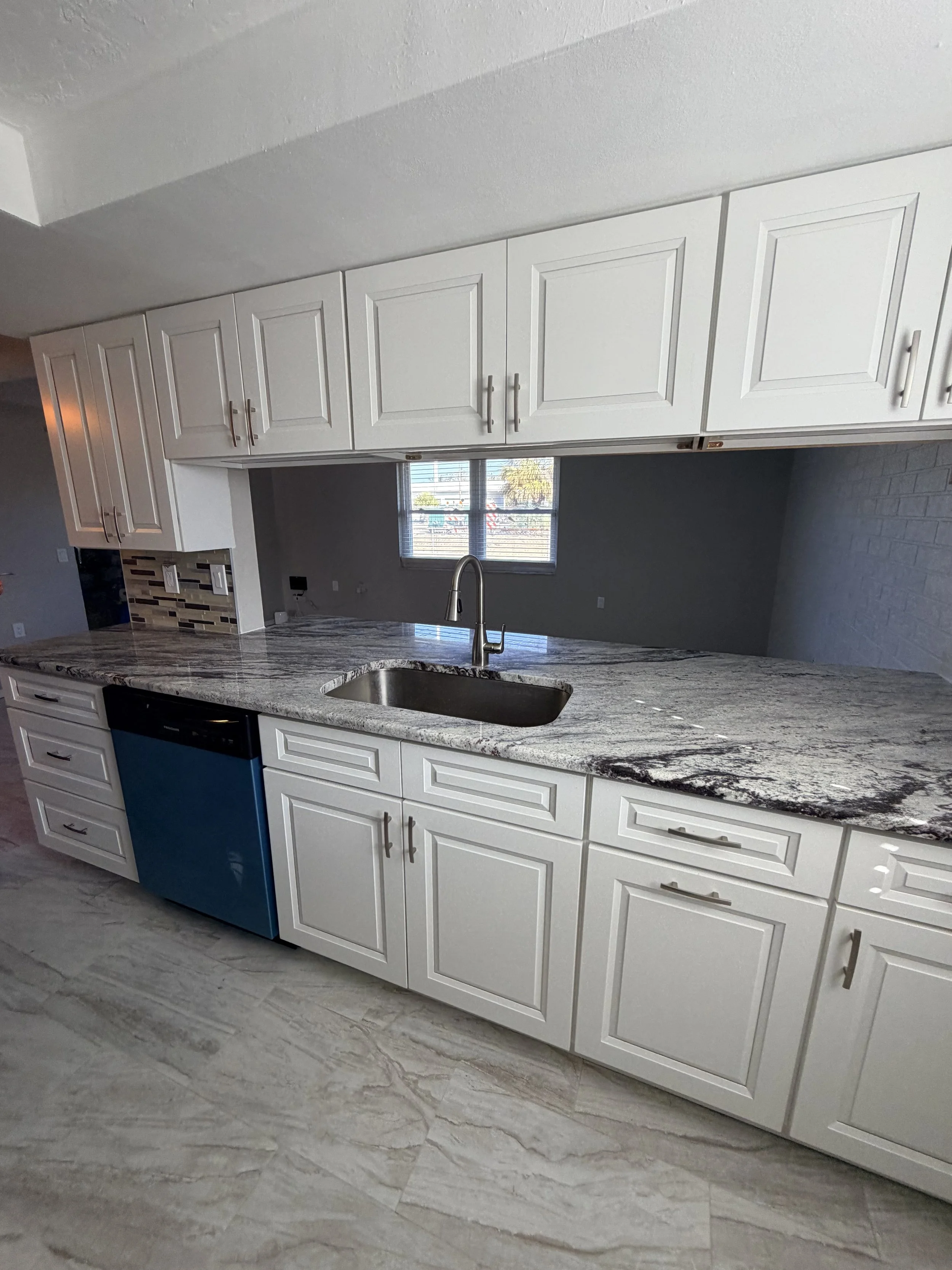 White kitchen cabinets with silver handles, granite countertops, and a window showing outdoor scenery, with a partial view of a blue dishwasher and grey tiled floor.