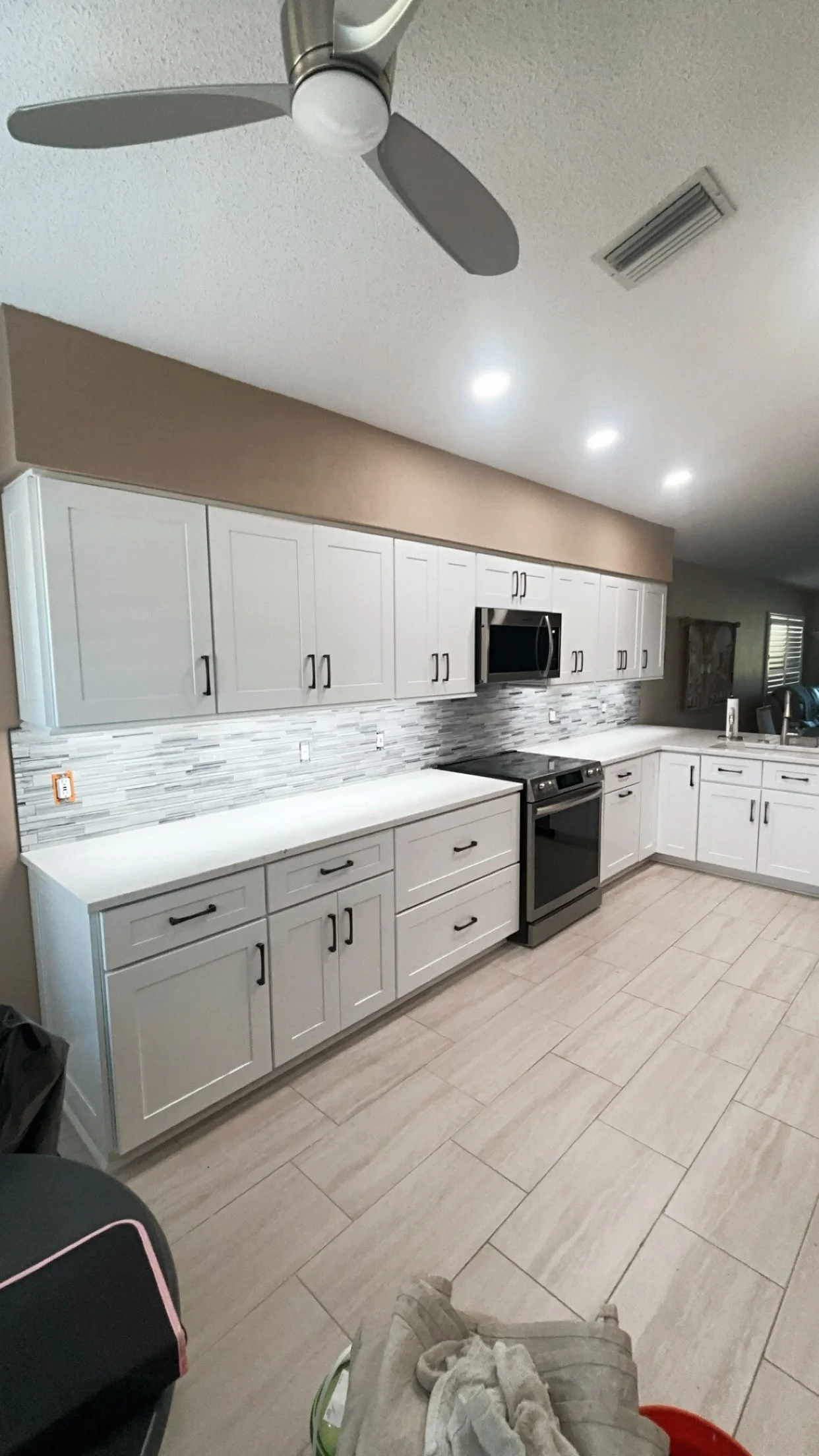 A modern kitchen with white cabinets, a gray tile backsplash, stainless steel appliances including a microwave and an oven, and a ceiling fan with three blades. The countertop is white, and the floor has beige tiles.