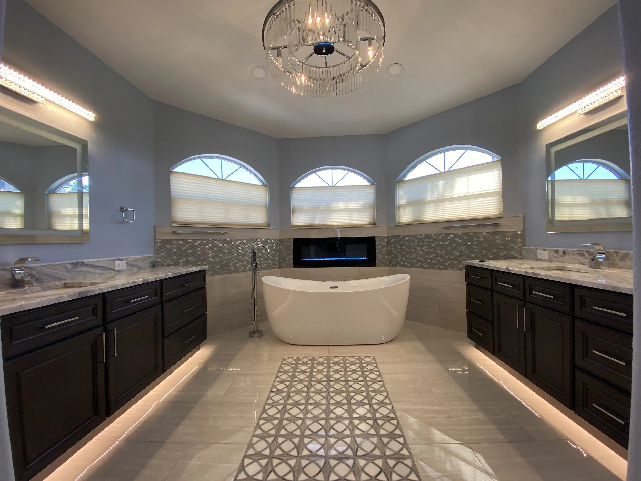 Modern bathroom with a freestanding bathtub, dark cabinets, marble countertops, large arched windows with blinds, a fireplace, a chandelier, and decorative tile flooring.